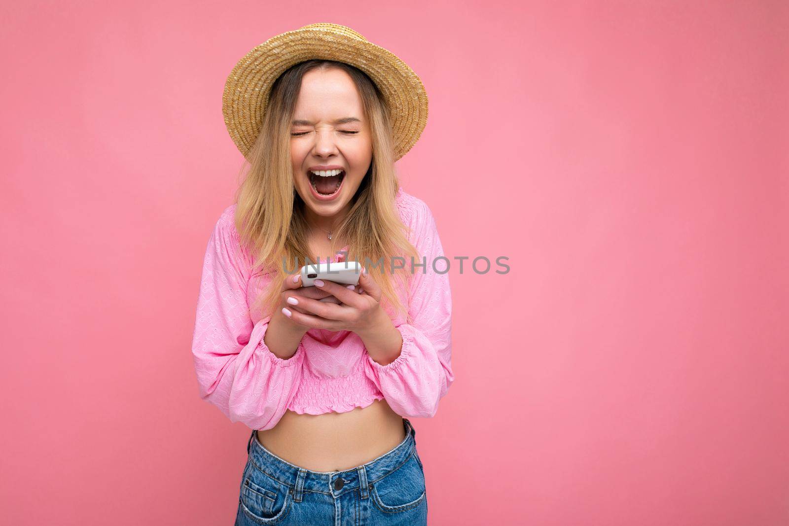 Attractive amazed young blonde woman smiling and having fun standing isolated over pink wall keeping hand in pocket wearing pink blouse and summer hat using mobile phone with close eyes.