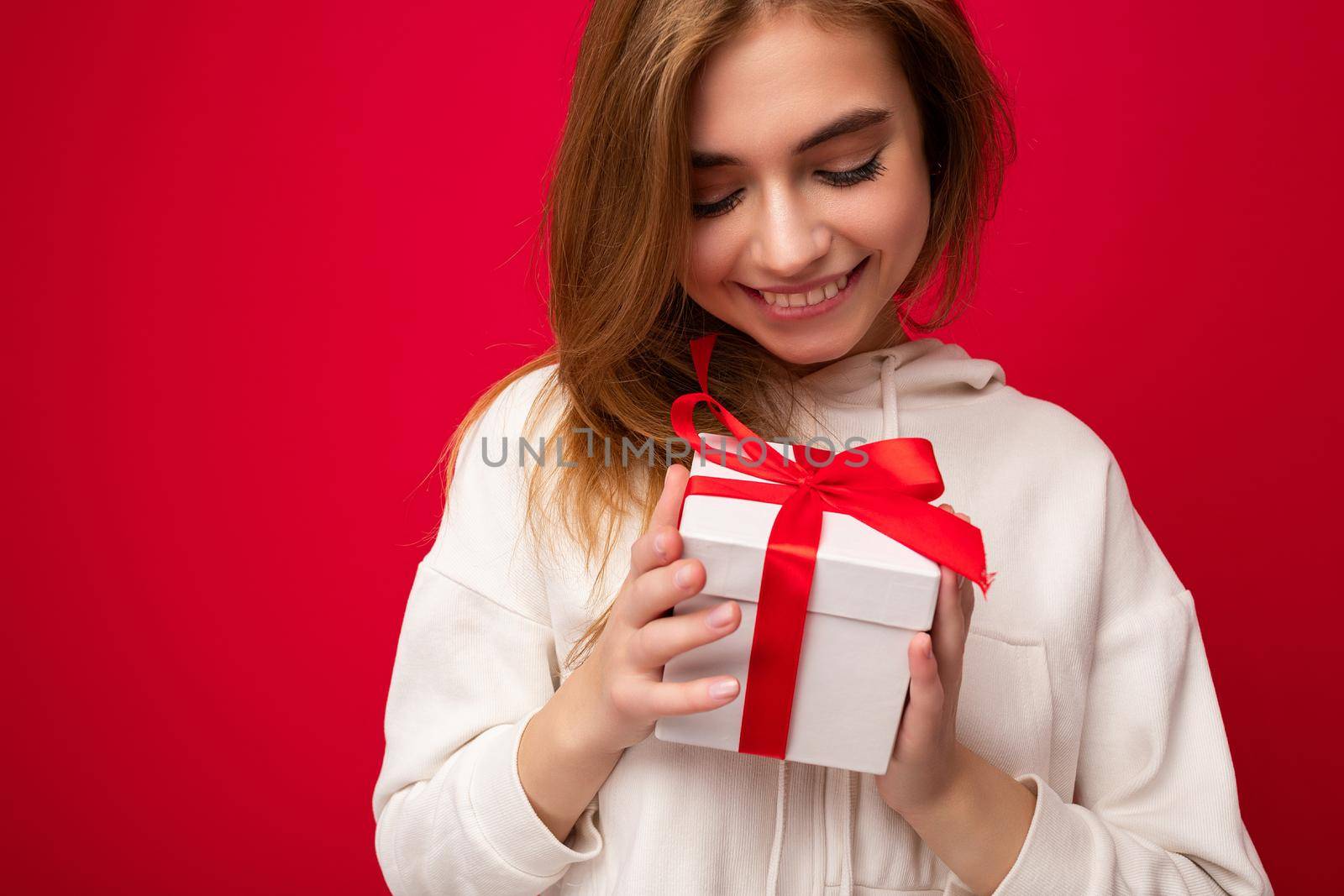 Photo shot of pretty positive smiling young dark blond woman isolated over colourful background wall wearing trendy outfit look holding gift box and looking at present box with red ribbon.