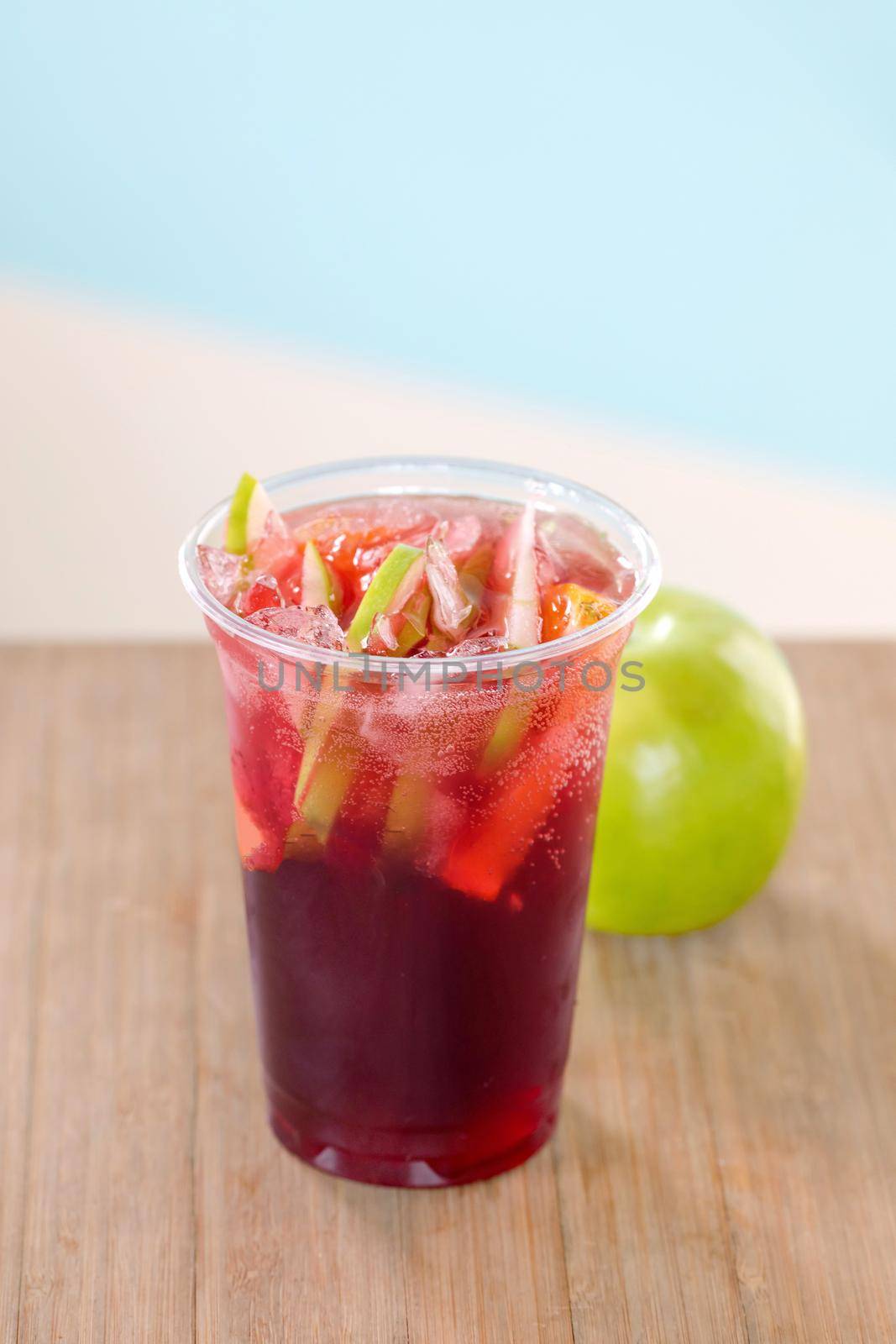 Red sangria beverage with fruits and ice in plastic glass standing on wooden table with green apple