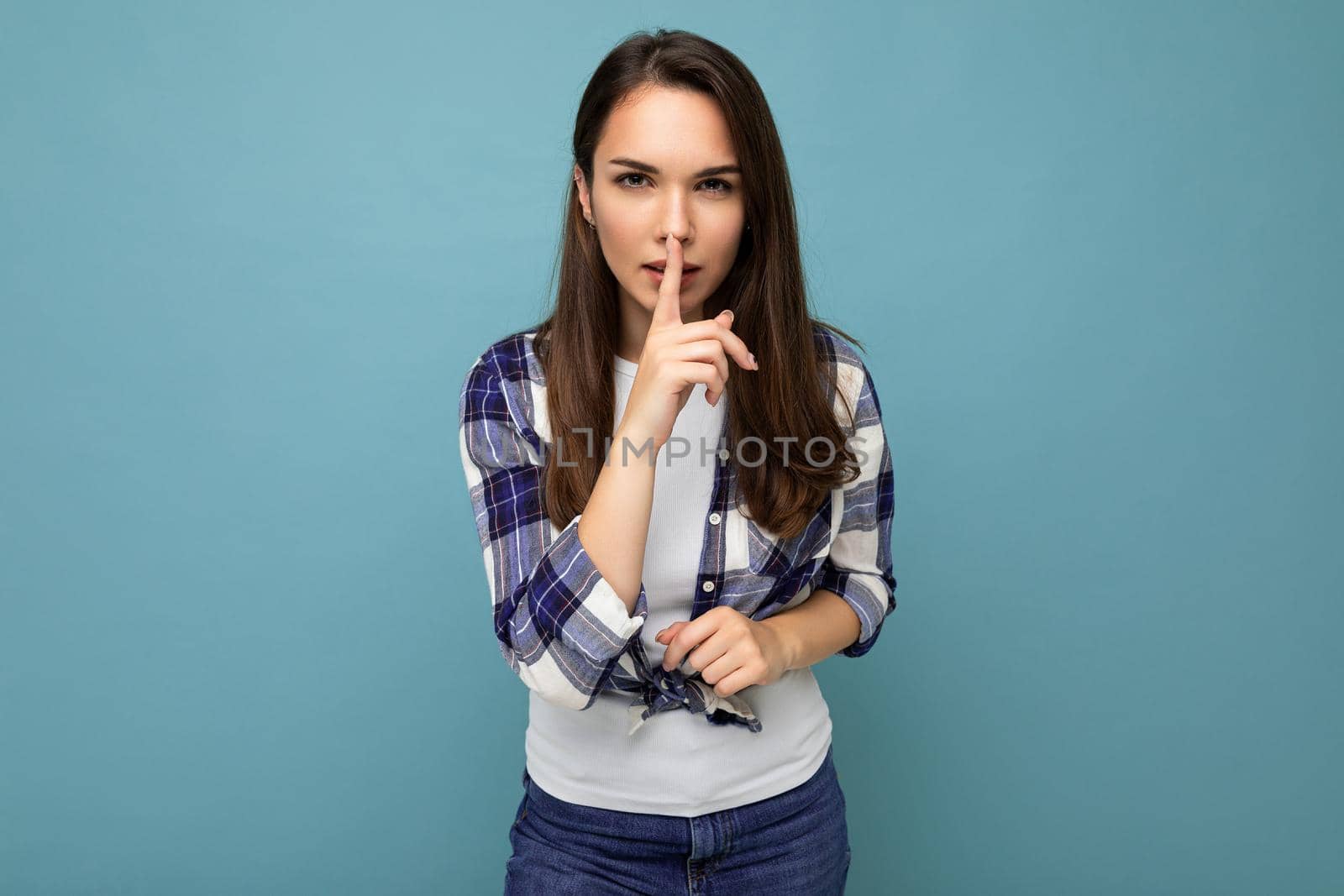 Young serious beautiful brunet woman with sincere emotions wearing trendy check shirt standing isolated on blue background with empty space and showing shhh gesture. Keep secret concept.