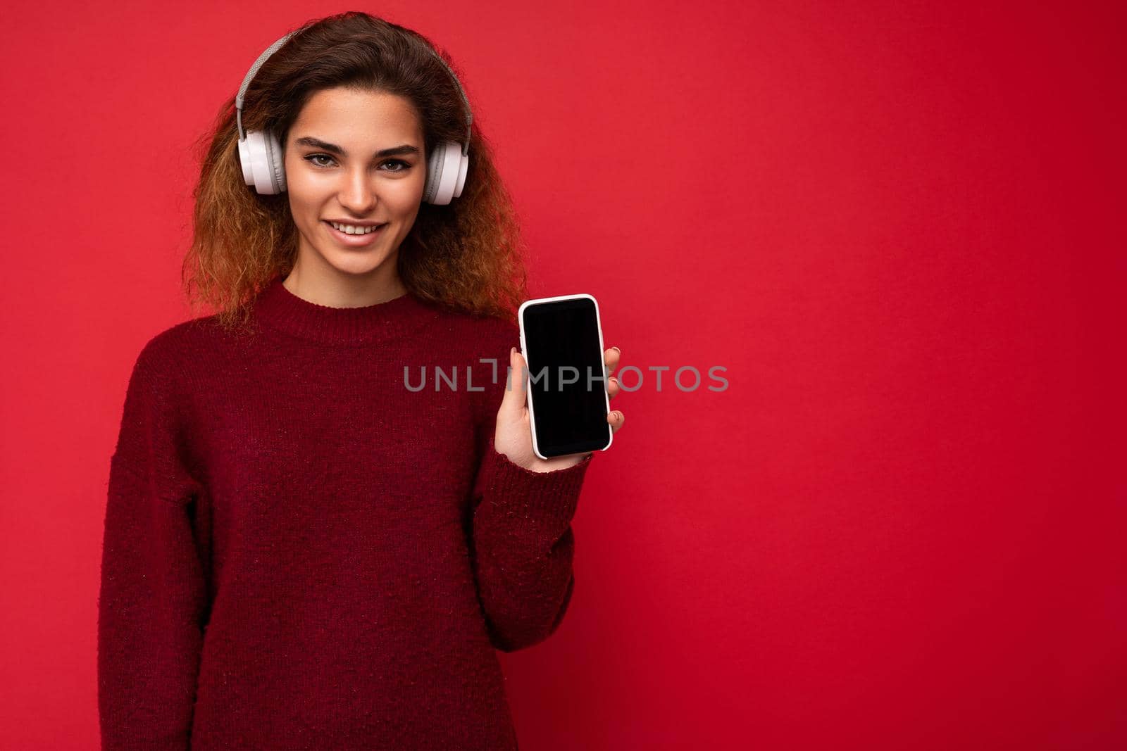 Beautiful happy smiling young woman wearing stylish casual outfit isolated on background wall holding and showing mobile phone with empty display for mockup wearing white bluetooth headphones listening to music and having fun looking at camera.