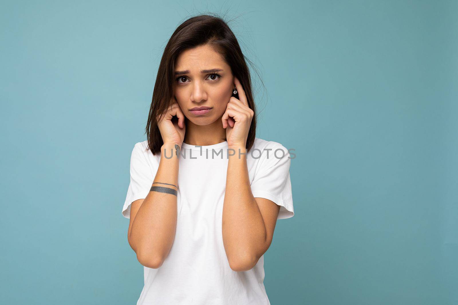 Portrait of sad sorrowful young pretty nice brunette woman with sincere emotions wearing casual white t-shirt for mockup isolated on blue background with copy space and covering ears with fingers.