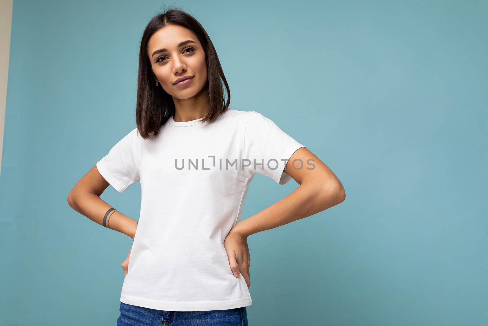 Photo of positive young beautiful brunette woman with sincere emotions wearing casual white t-shirt for mockup isolated over blue background with copy space.