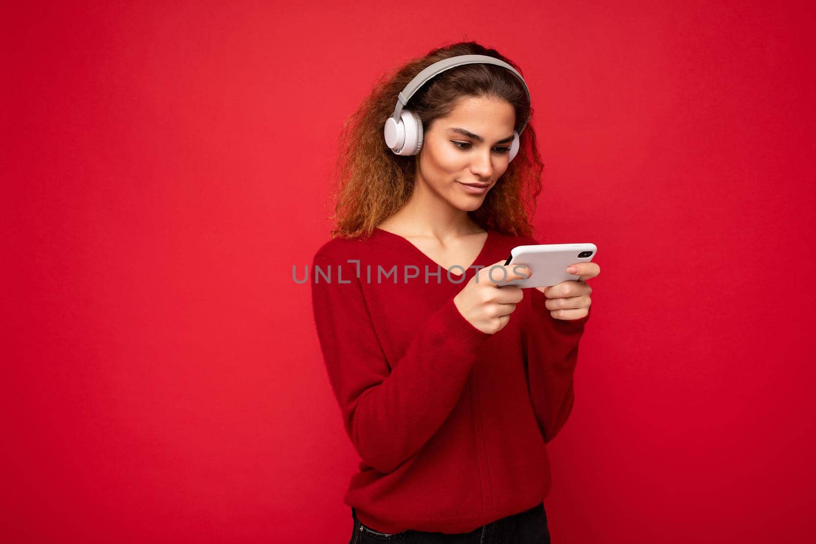 Photo shot of beautiful joyful smiling young female person wearing stylish casual outfit isolated over colorful background wall wearing white bluetooth wireless earphones and listening to music and using mobile phone looking at gadjet display.