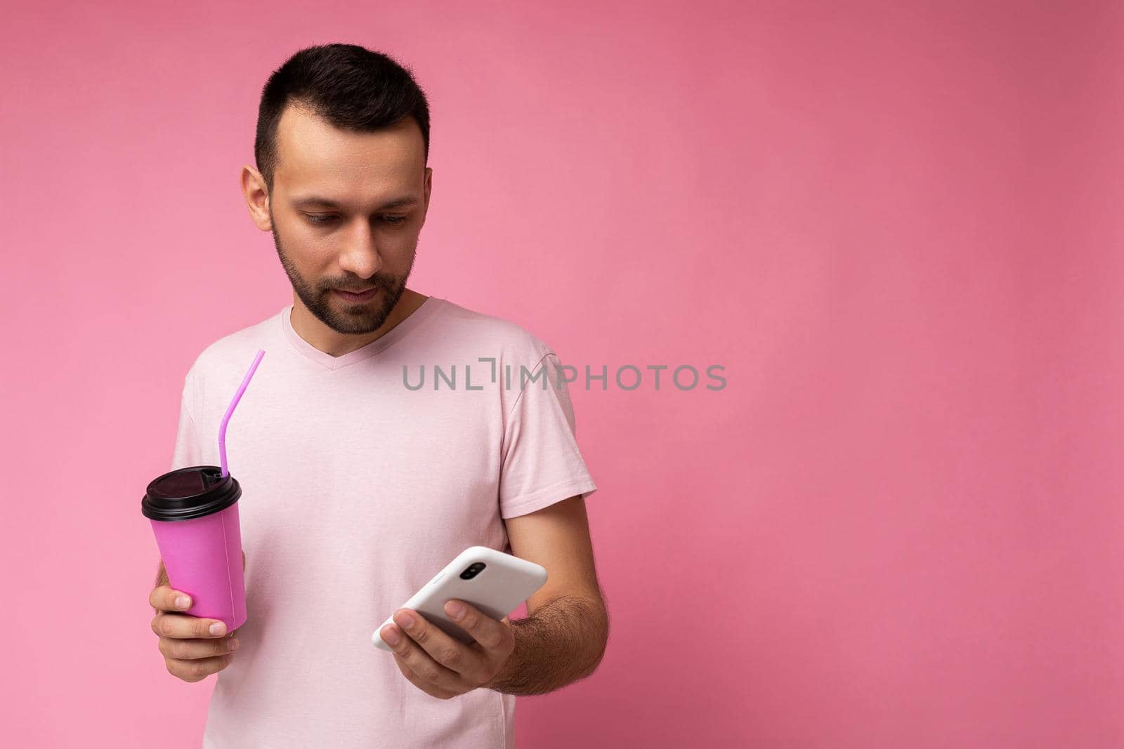 Photo shot of handsome positive good looking young man wearing casual stylish outfit poising isolated on background with empty space holding in hand and using mobile phone messaging sms looking at smartphone display screen.