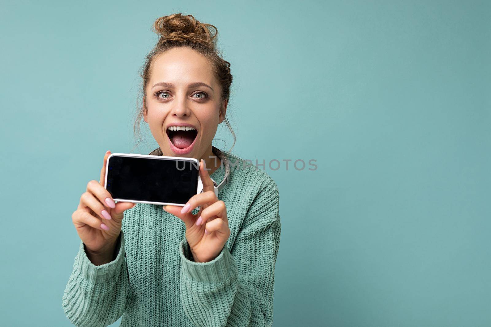 Photo of amazed shocked beautiful young woman good looking wearing casual outfit standing isolated on background with copy space holding smartphone showing phone in hand with empty screen display for mockup looking at camera.
