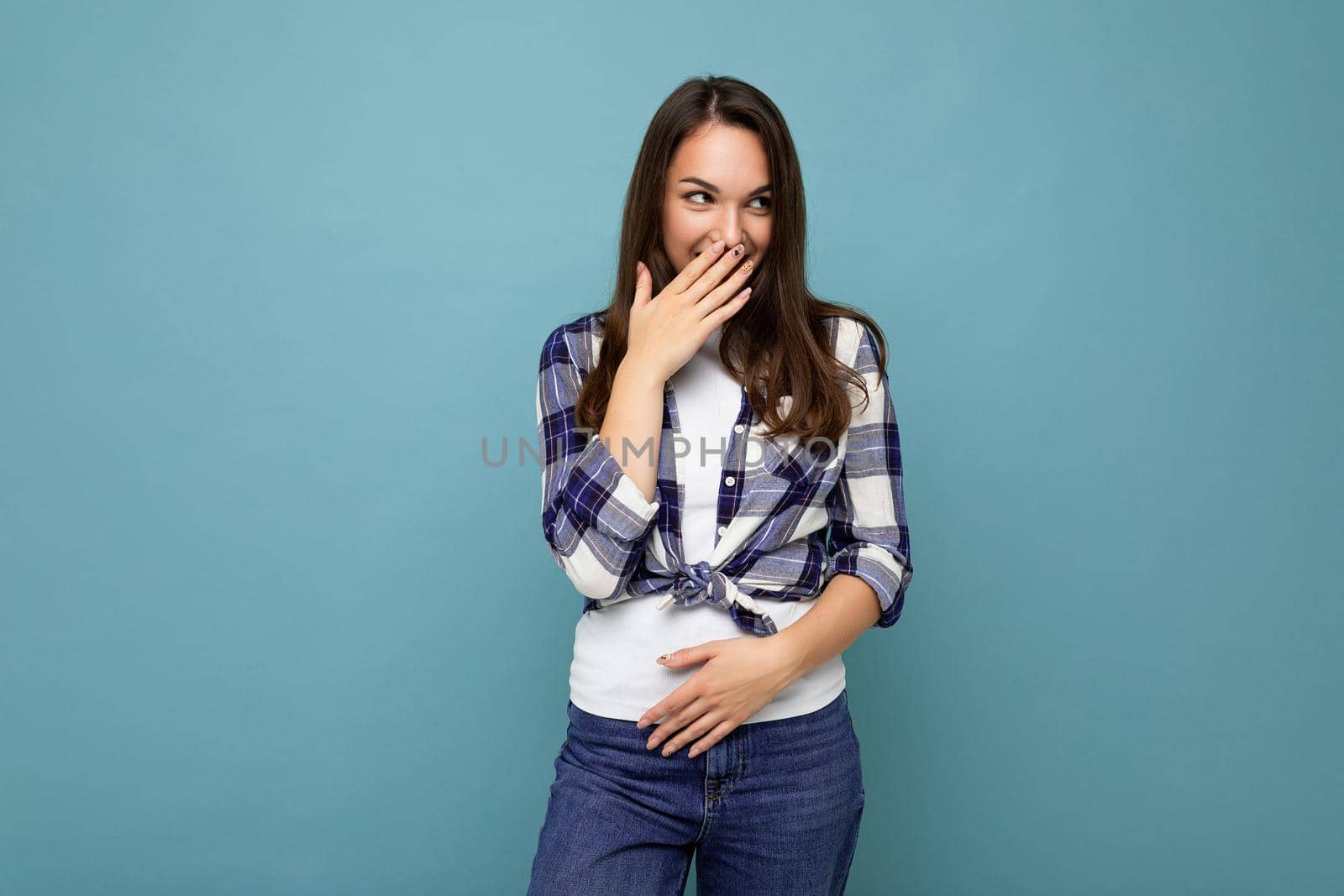 Young positive happy attractive pretty brunet woman with sincere emotions wearing stylish check shirt standing isolated on blue background with empty space and laughing.