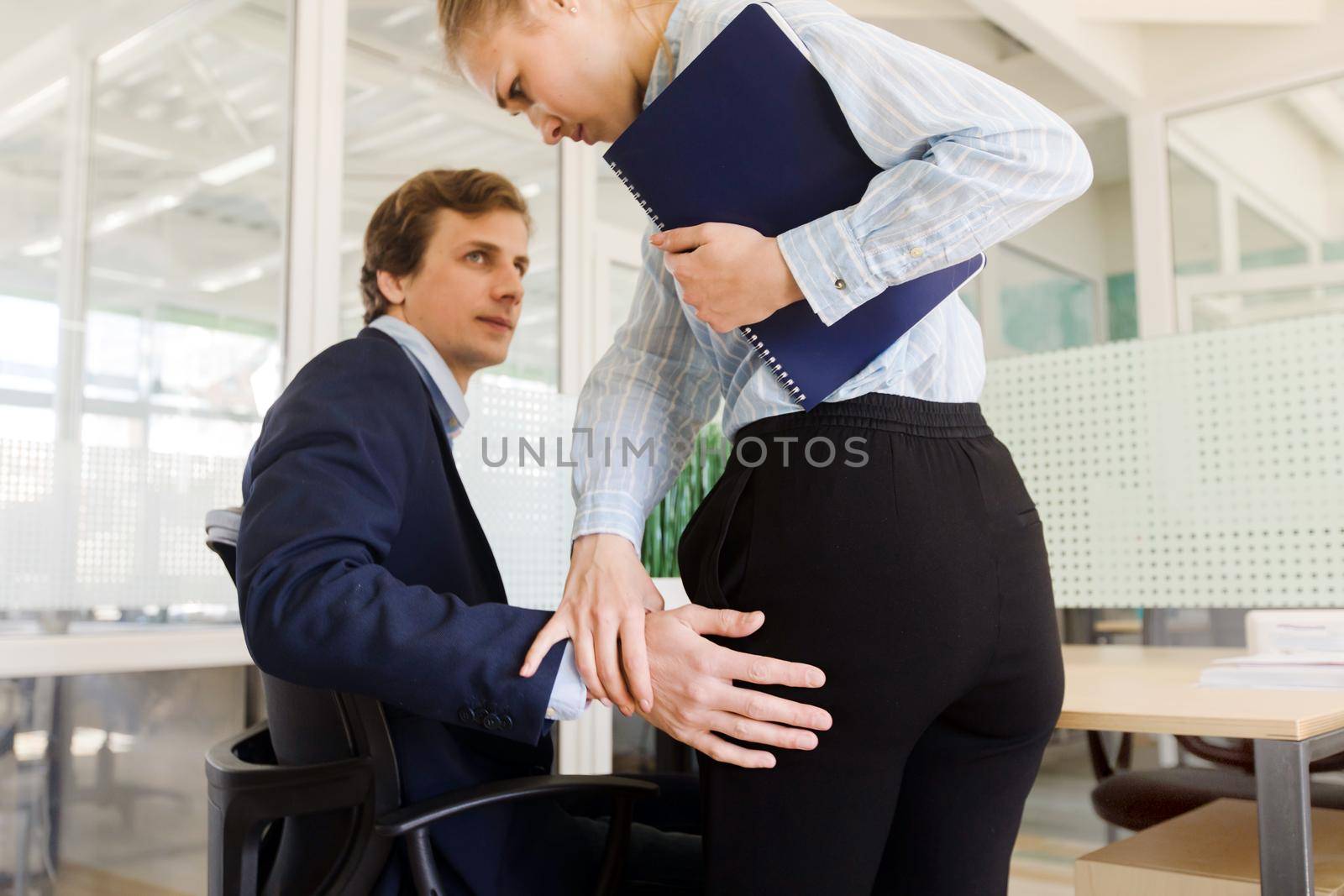 Young formal businessman sitting at table in office and touching hips of woman in sexual harassment