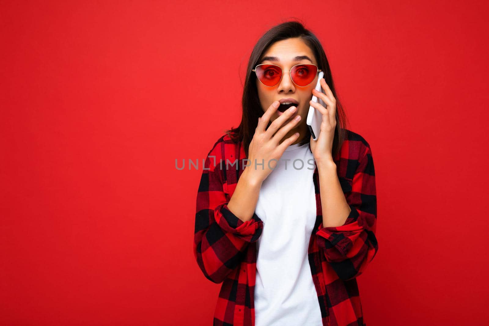 Beautiful shocked amazed young brunette woman wearing stylish red shirt white t-shirt and red sunglasses isolated over red background talking on mobile phone looking at camera.