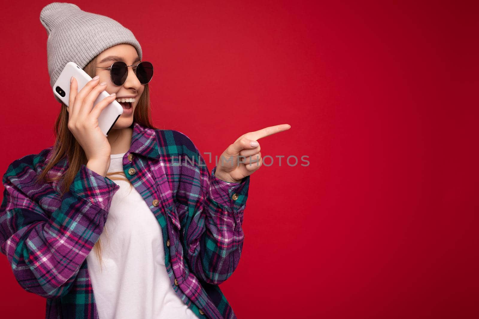 photo of beautiful happy positive young blonde woman wearing hipster purple shirt and casual white t-shirt grey hat and sunglasses isolated over red background holding in hand and talking on mobile phone looking to the side and pointing finger at empty space by TRMK