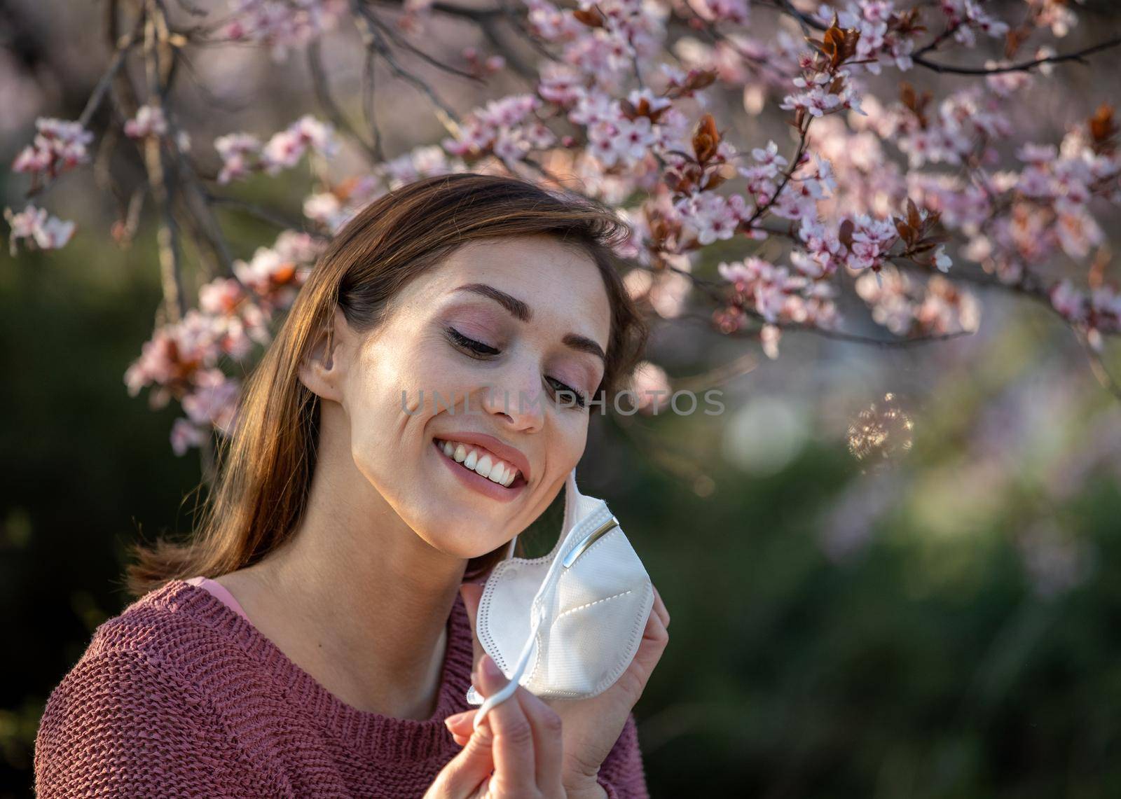 Smiling young woman putting facial mask on and looking at blooming tree. Spring allergy attack protection