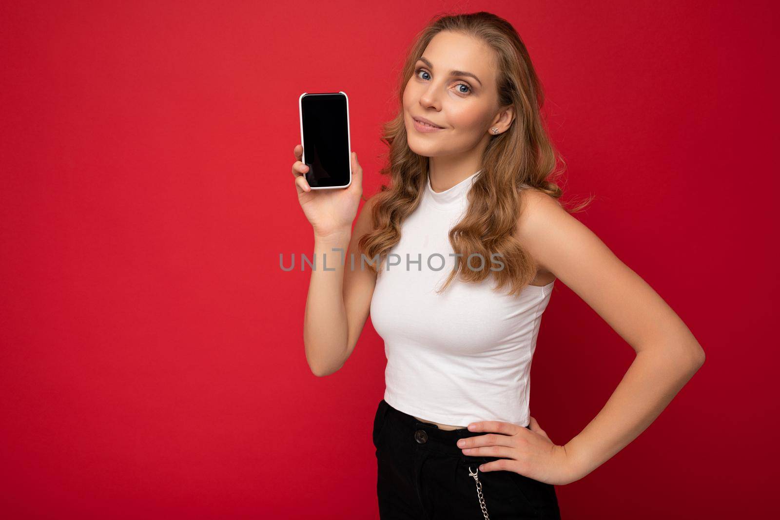 beautiful young blonde woman wearing white t-shirt isolated on red background with copy space holding smartphone showing phone in hand with empty screen for mockup looking at camera by TRMK