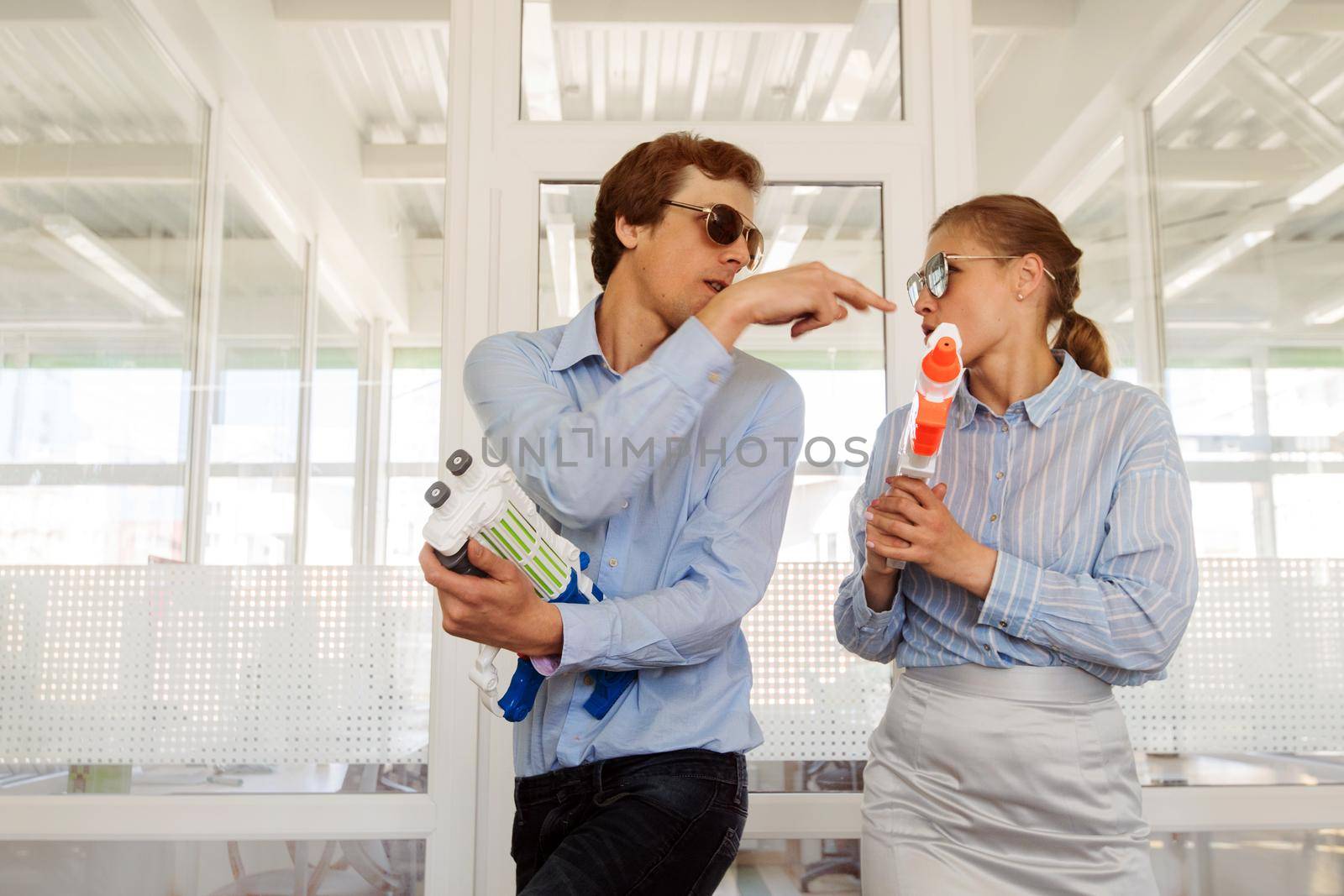 Stylish modern woman and man in sunglasses having fun in office with toy plastic guns
