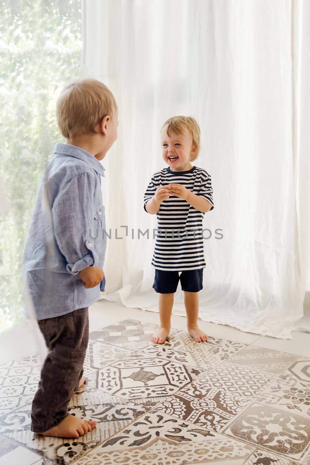 Adorable toddler with little boy standing indoors near window and laughing cheerfully looking at each other
