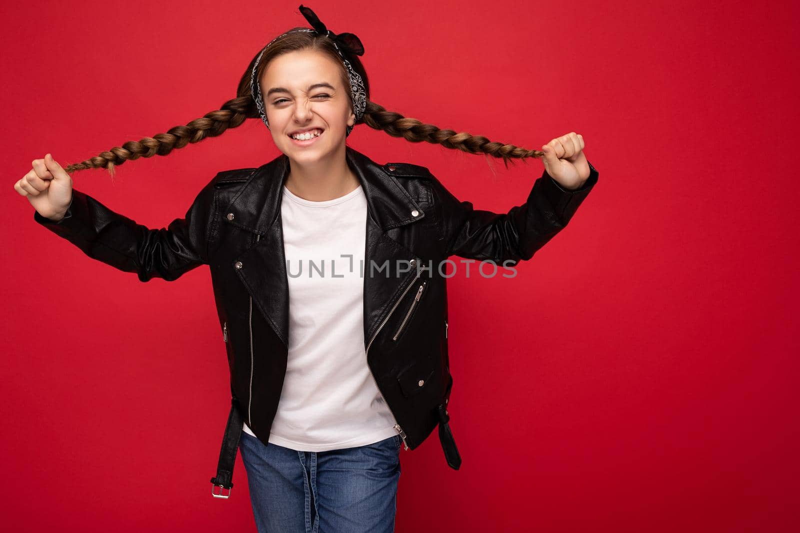 Photo shot of beautiful happy smiling brunette little girl with pigtails wearing trendy black leather jacket and white t-shirt for mockup standing isolated over red background wall looking at camera.