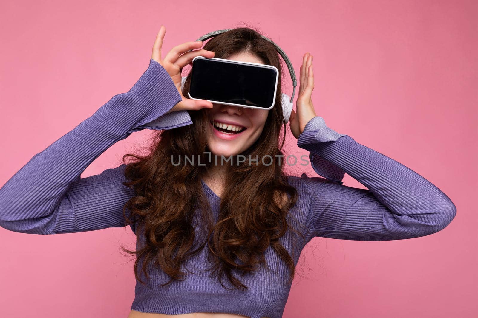 Photo of pretty positive smiling young brunette curly woman wearing purple crop top isolated over pink background wall holding and suggesting mobile phone with empty display and bluetooth headsets.