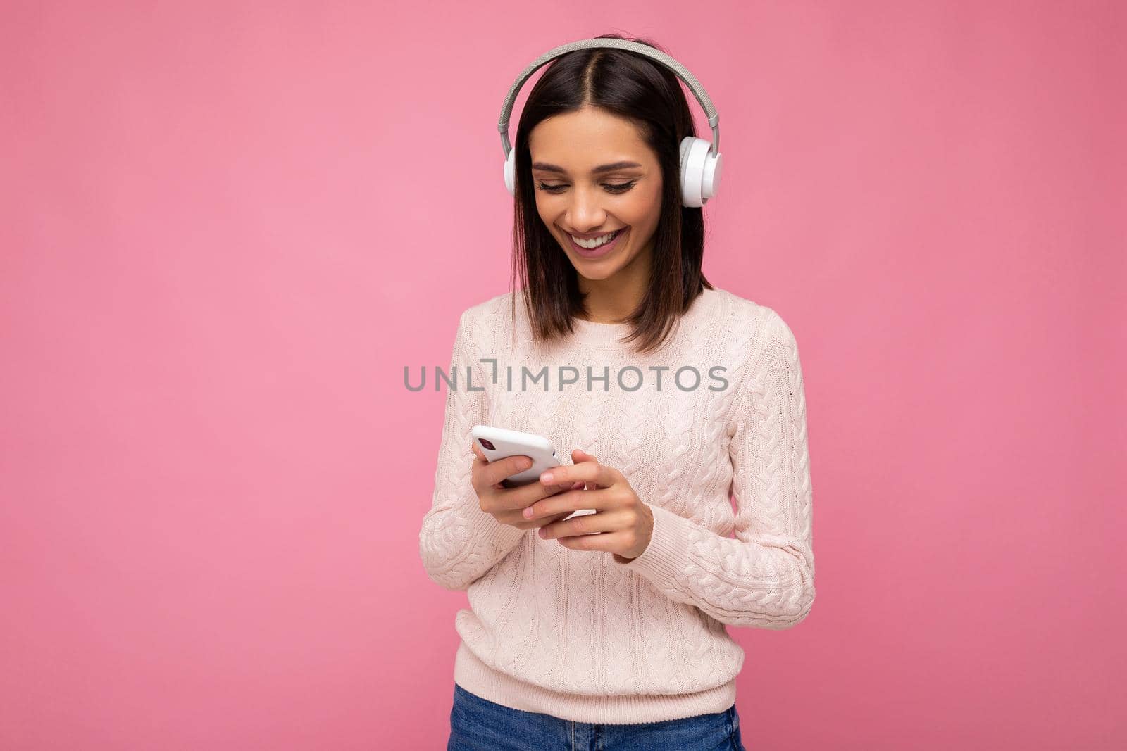 Photo shot of beautiful joyful smiling young female person wearing stylish casual outfit isolated over colorful background wall wearing white bluetooth wireless earphones and listening to music and using mobile phone looking at gadjet display.