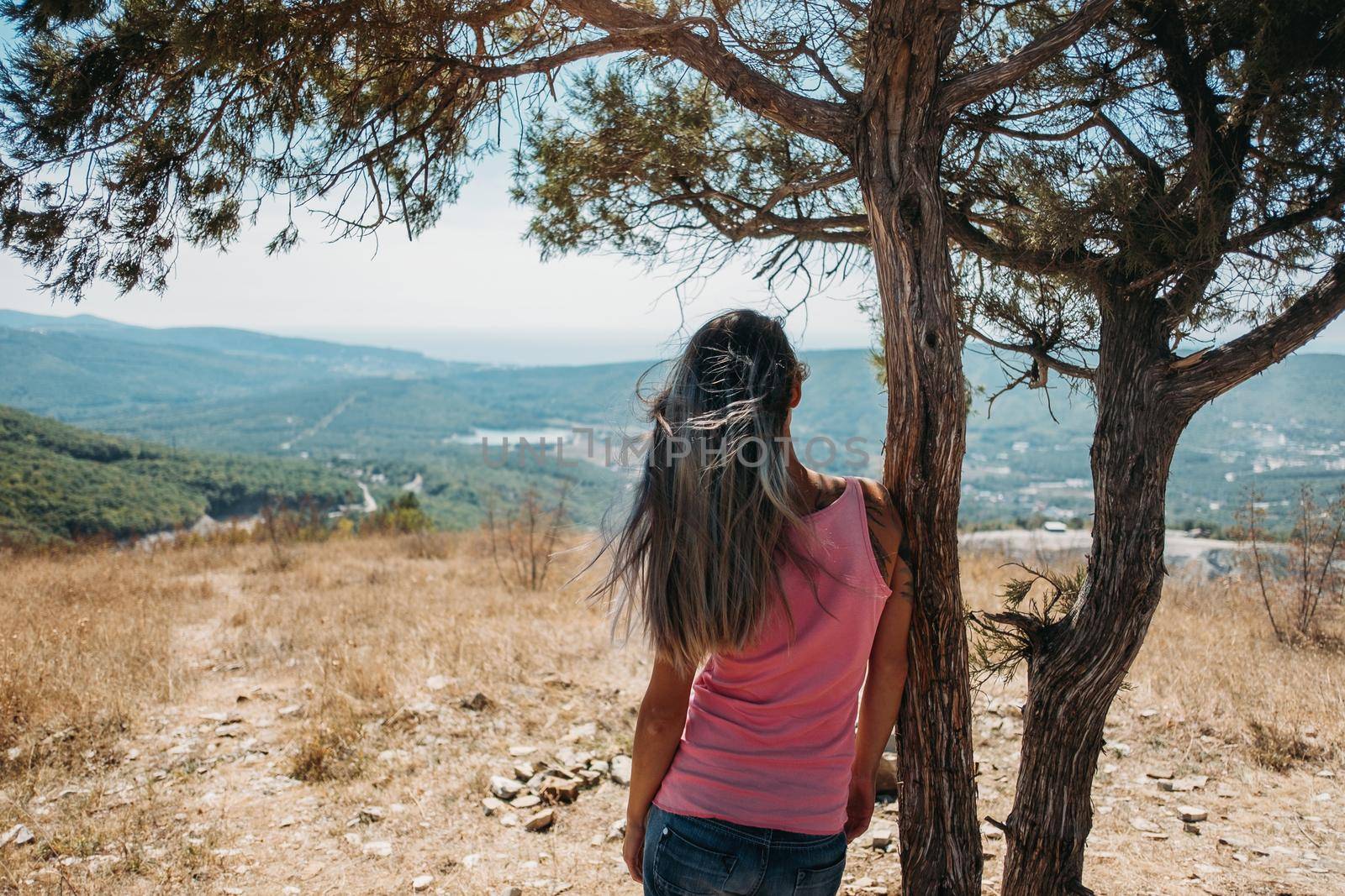 Young woman standing near a tree and looking at town in the mountains in summer, rear view