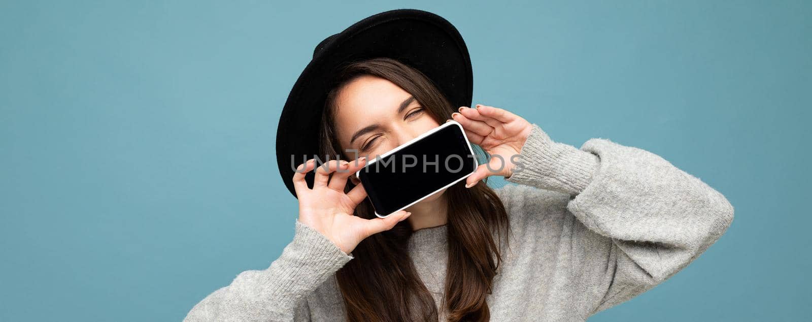 panoramic Photo of Beautiful positive woman person wearing black hat and grey sweater holding mobilephone showing smartphone isolated on background with fantastic eyes.Mock up, cutout, free space
