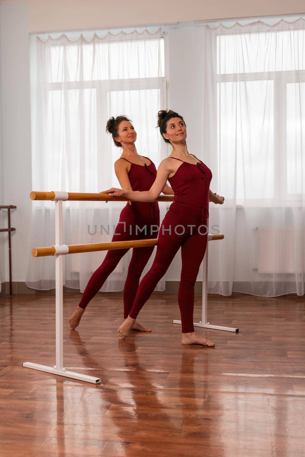 Two young womans fitness instructor in red Sportswear Leggings and Top stretching in the gym before pilates, on a yoga mat near the large window on a sunny day, female fitness yoga routine concept by panophotograph