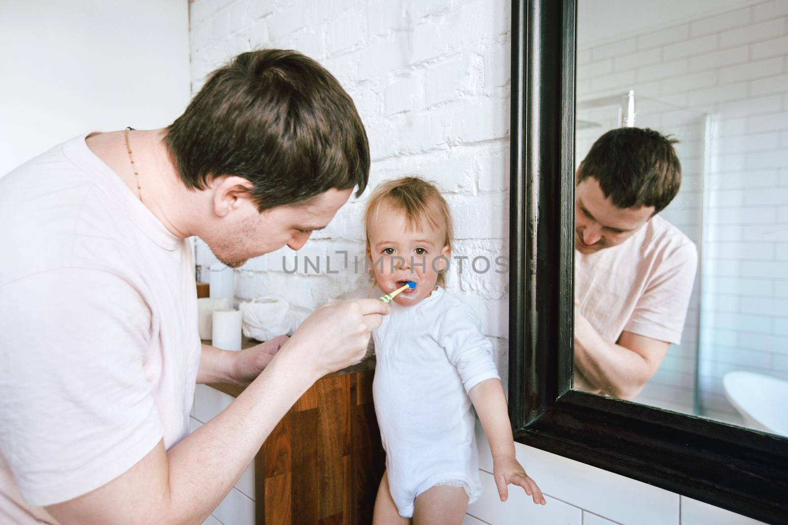 Happy father helping with teeth brushing to charming little boy in light white bathroom