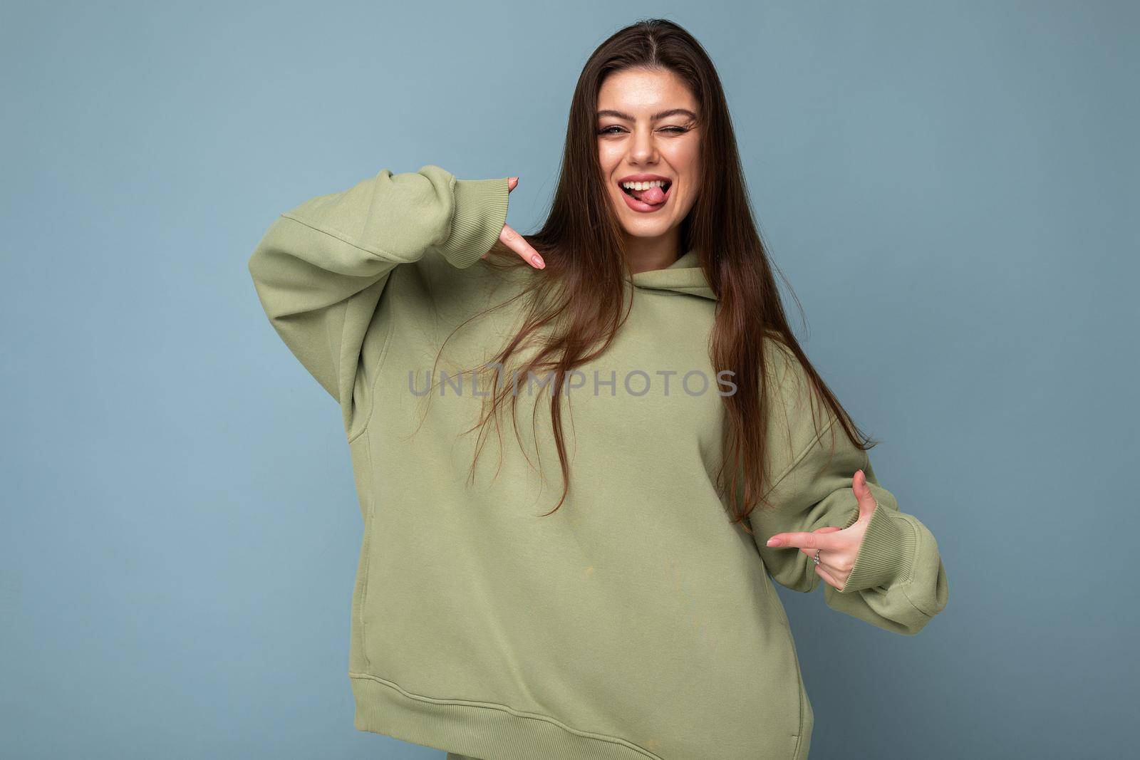 Photo of beautiful positive joyful young brunette cool woman in a stylish khaki hoodie isolated on blue background. Mockup. Empty space.