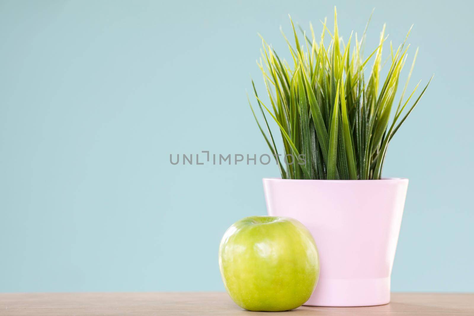 Delicious green apple lying on light blue background near ceramic pot with nice plant
