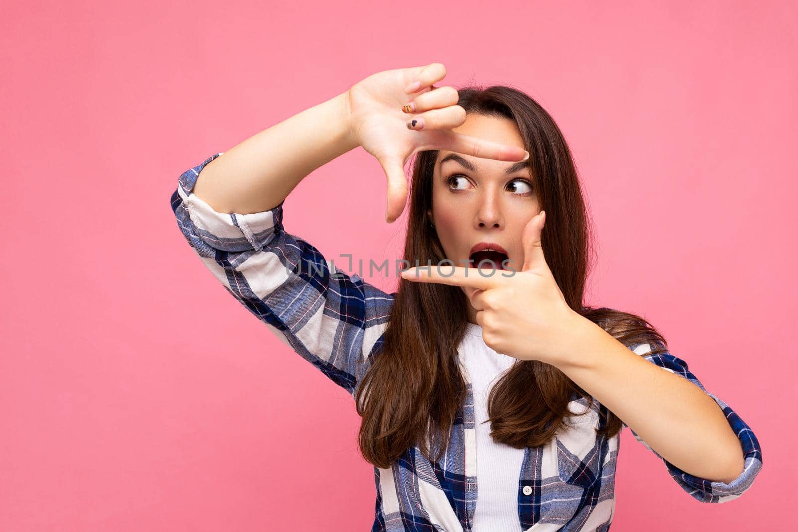 Young positive delightful amazed pretty brunette woman with sincere emotions wearing hipster check shirt making frame with hands, taking picture with imaginary camera and standing isolated on pink background with copy space.