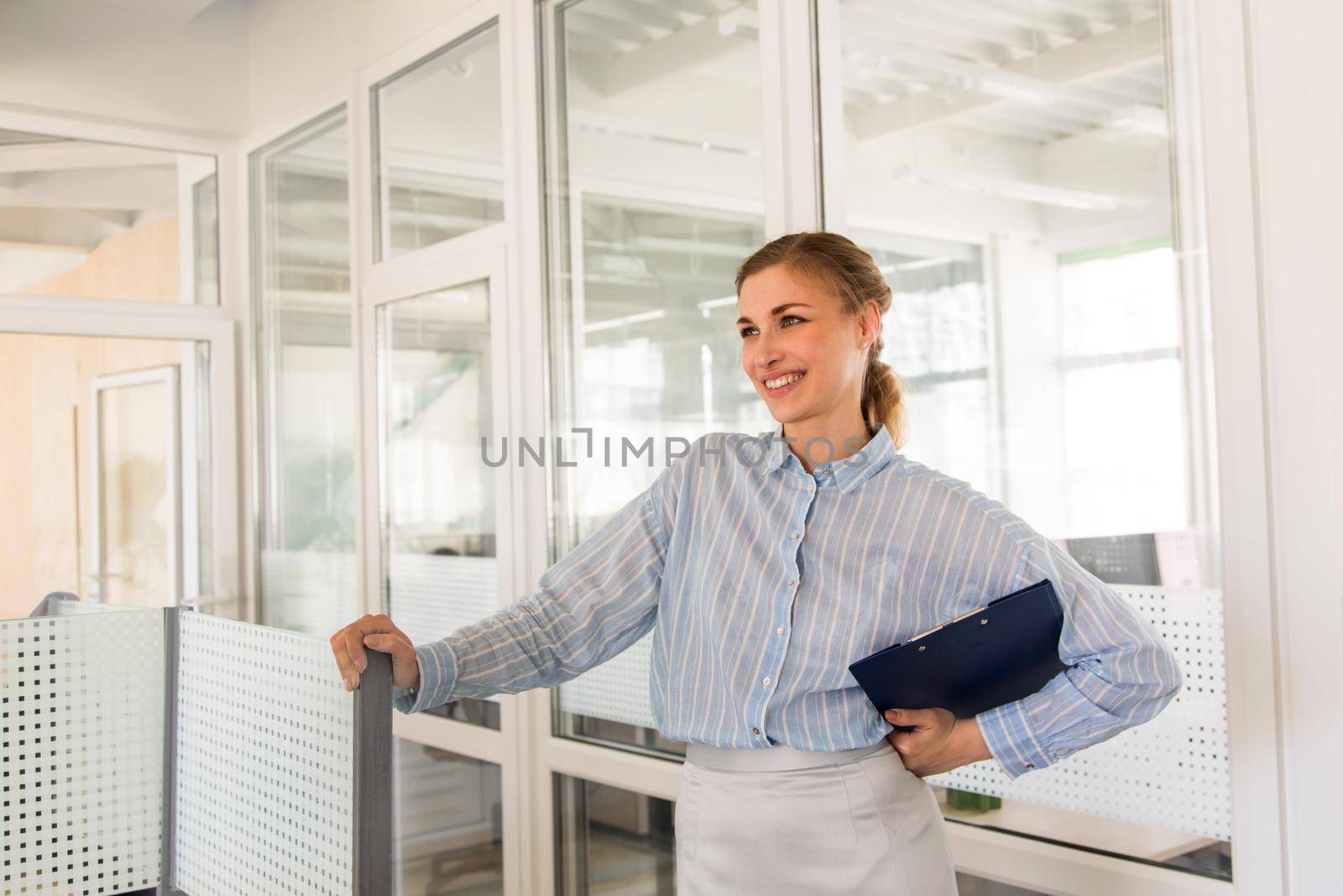 Pretty young female in formal wear holding clipboard and smiling while standing in office and leaning on partition