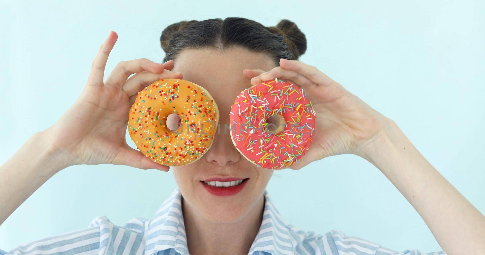 Funny portrait of young attractive brunette on bright turquoise backgroung is taking two round donuts and makes sunglasses from it, smiling and laughing