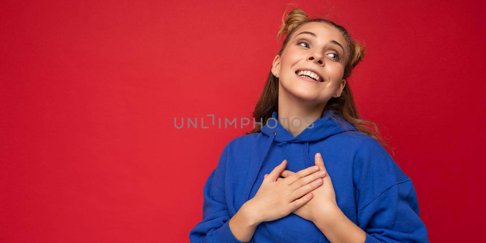 Panoramic portrait of young happy smiling positive pretty cute blonde woman with two horns with sincere emotions wearing hipster blue hoodie isolated on red background with free space and holding hands on chest by TRMK