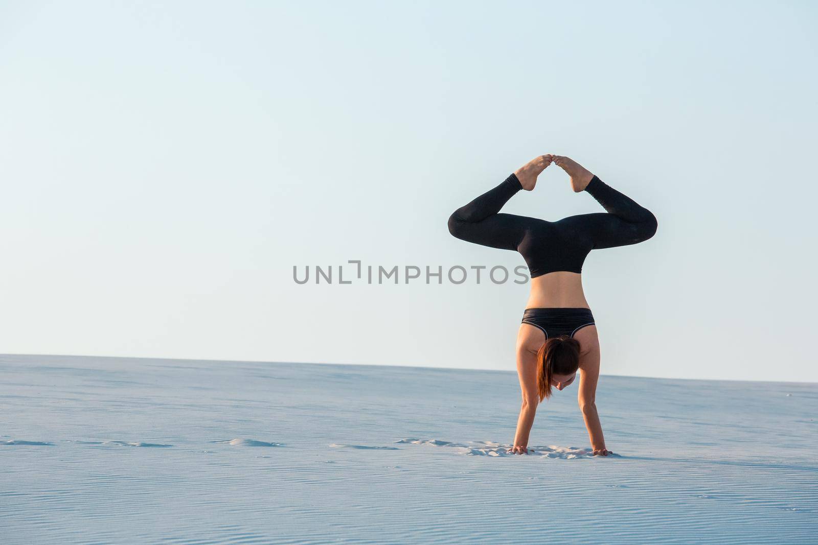 Young woman practicing inversion balancing yoga pose handstand on sand. Sunset and white sand.