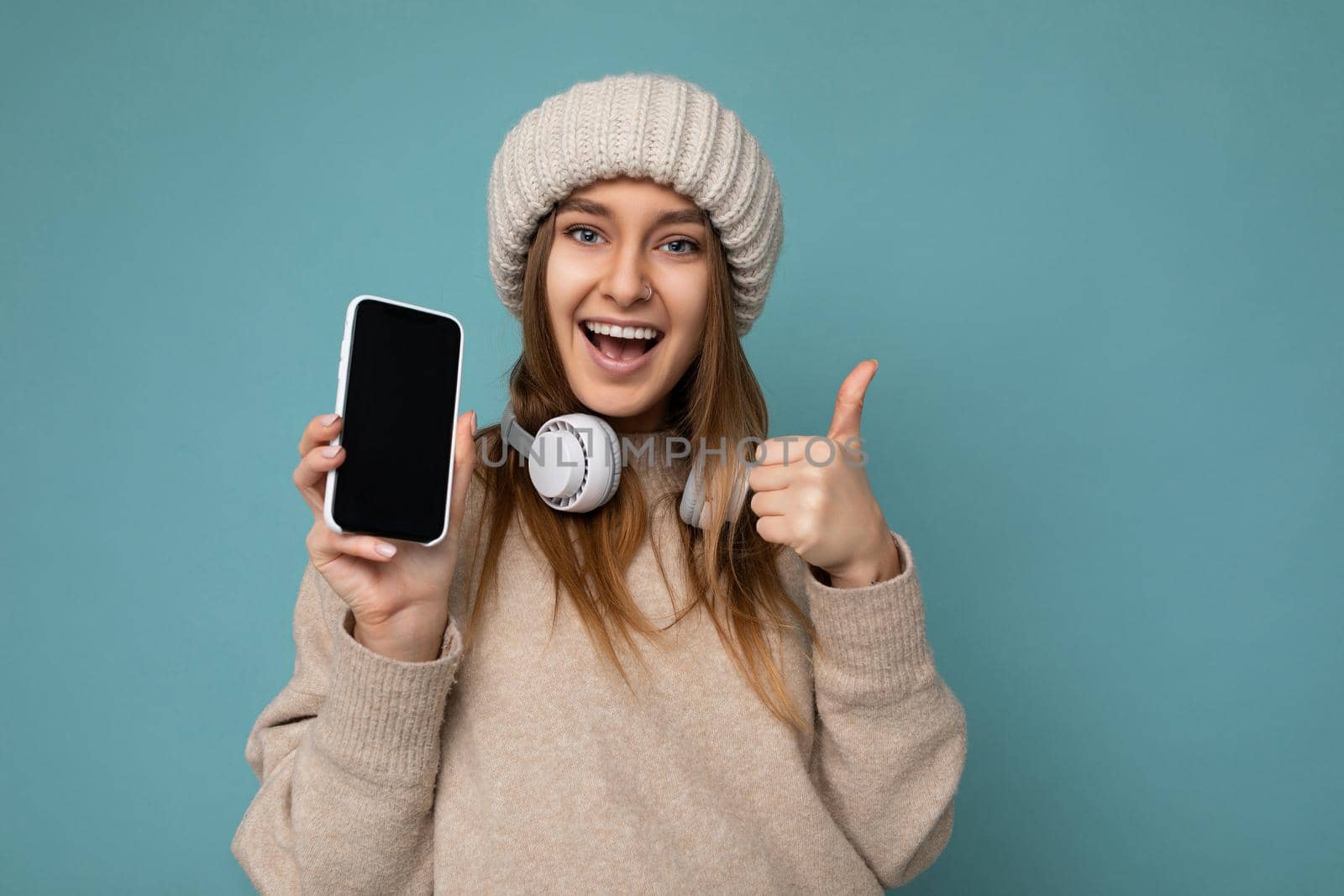 Photo of attractive positive joyful young dark blonde woman wearing beige stylish sweater and knitted beige winter hat isolated on blue background wearing white bluetooth wireless earphones and listening to cool music and showing mobile phone with empty display for mockup looking at camera and showing thumbs up gesture.