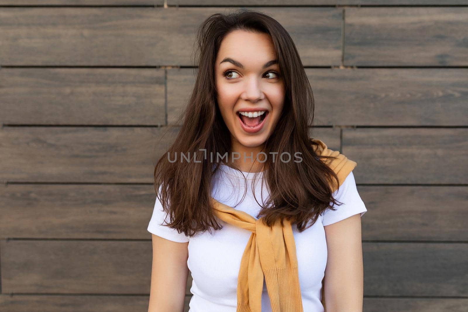 Portrait of successful smiling joyful happy young brunet woman wearing casual white t-shirt and jeans with yellow sweater poising near brown wall in the street and having fun.