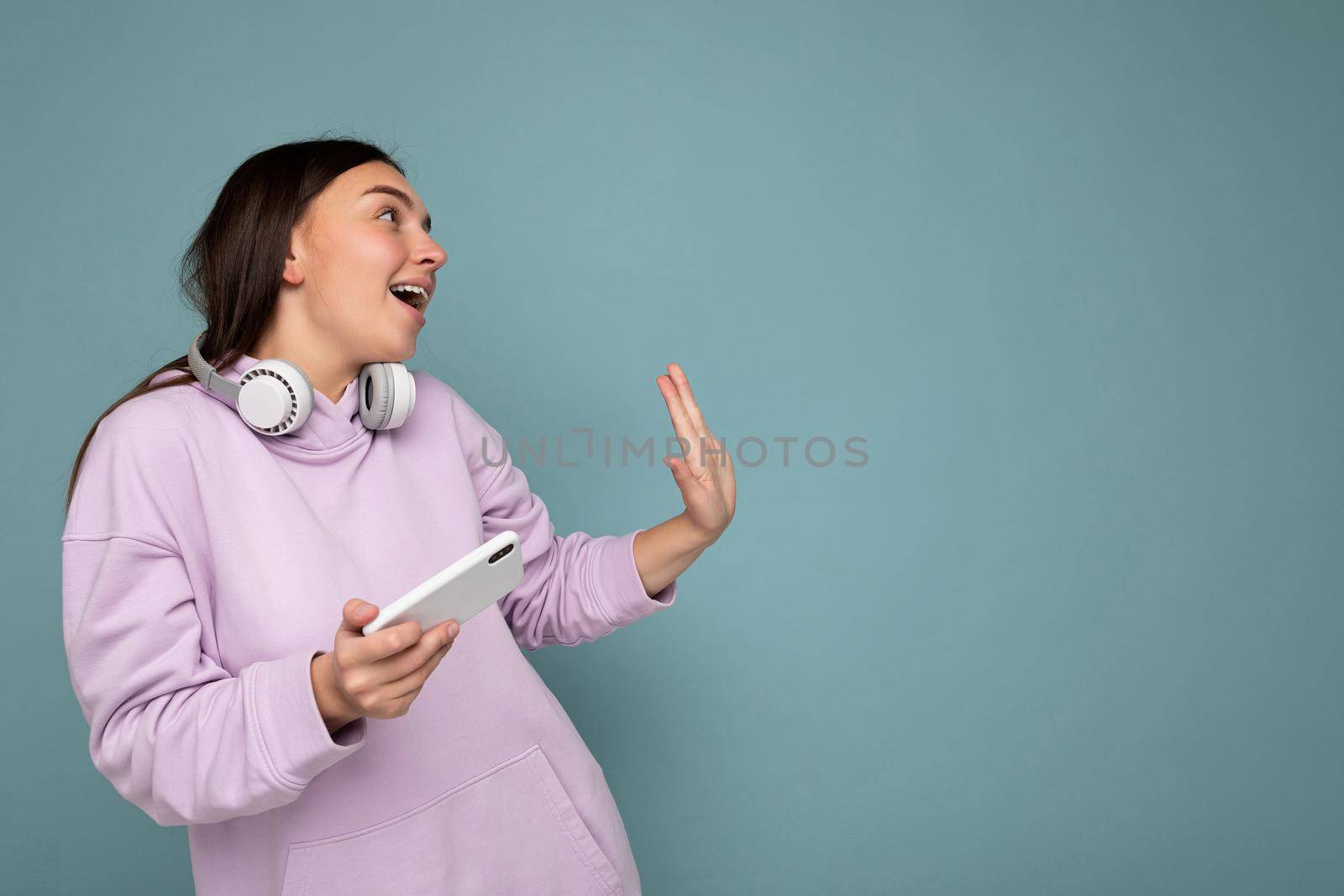 Beautiful happy smiling young brunette woman wearing purple hoodie isolated on blue background holding and using mobile phone surfing on the internet wearing white bluetooth headphones and having fun looking to the side and moving. Copy space