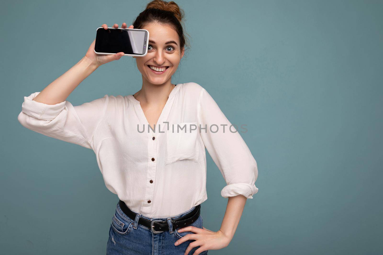 Photo of beautiful smiling young woman good looking wearing casual stylish outfit standing isolated on background with copy space holding smartphone showing phone in hand with empty screen display for mockup looking at camera.