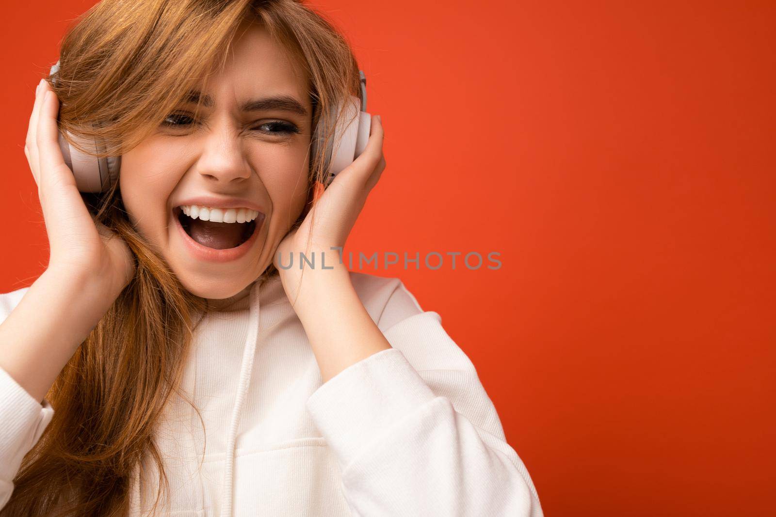 Portrait photo shot of attractive emotional positive young blonde woman wearing white hoodie isolated over colourful background wearing white wireless bluetooth headsets listening to music and having fun.