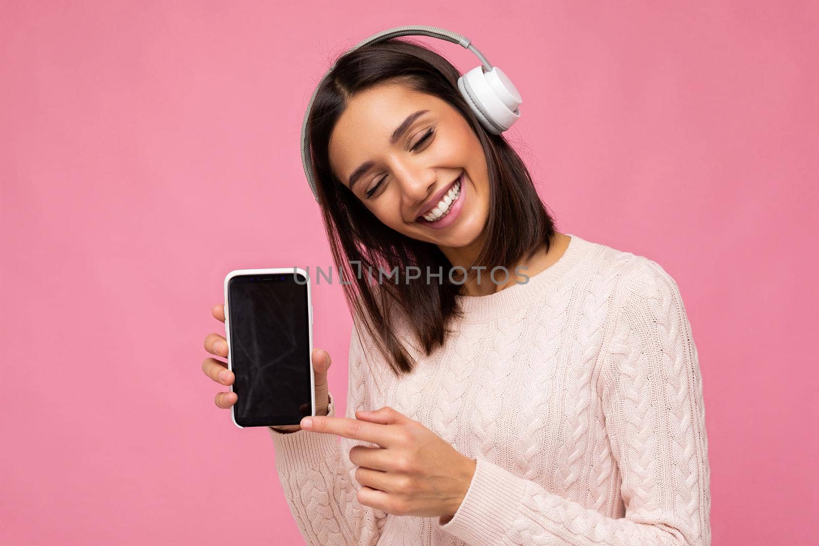 Beautiful cute young brunette woman wearing pink sweater isolated over pink background wall wearing white bluetooth wireless headphones and listening to good music and showing mobile phone with empty display for mockup having fun.