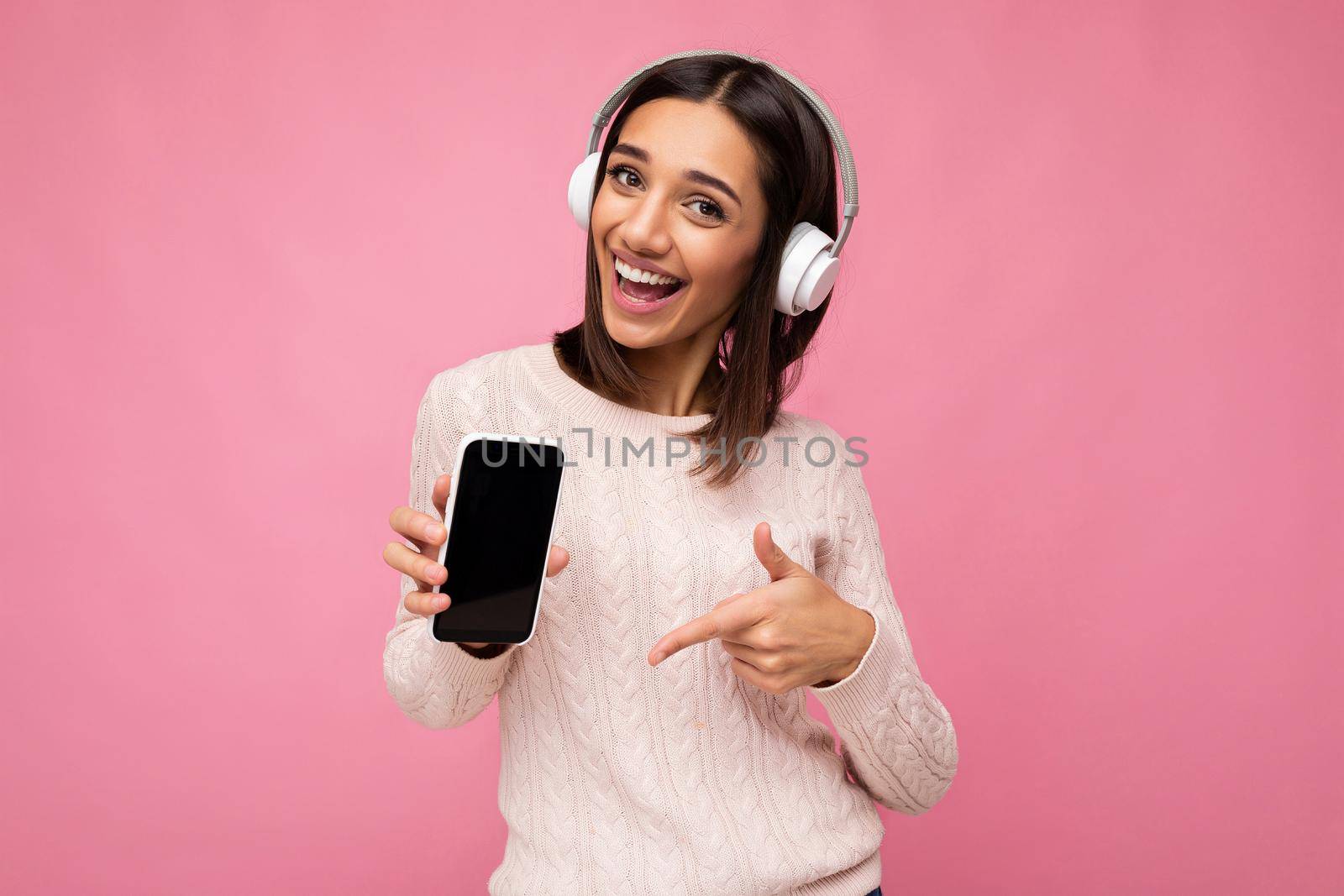 Attractive positive smiling young woman wearing stylish casual outfit isolated on colourful background wall holding and showing mobile phone with empty screen for cutout wearing white bluetooth headphones and having fun looking at camera and pointing finger at gadjet display.