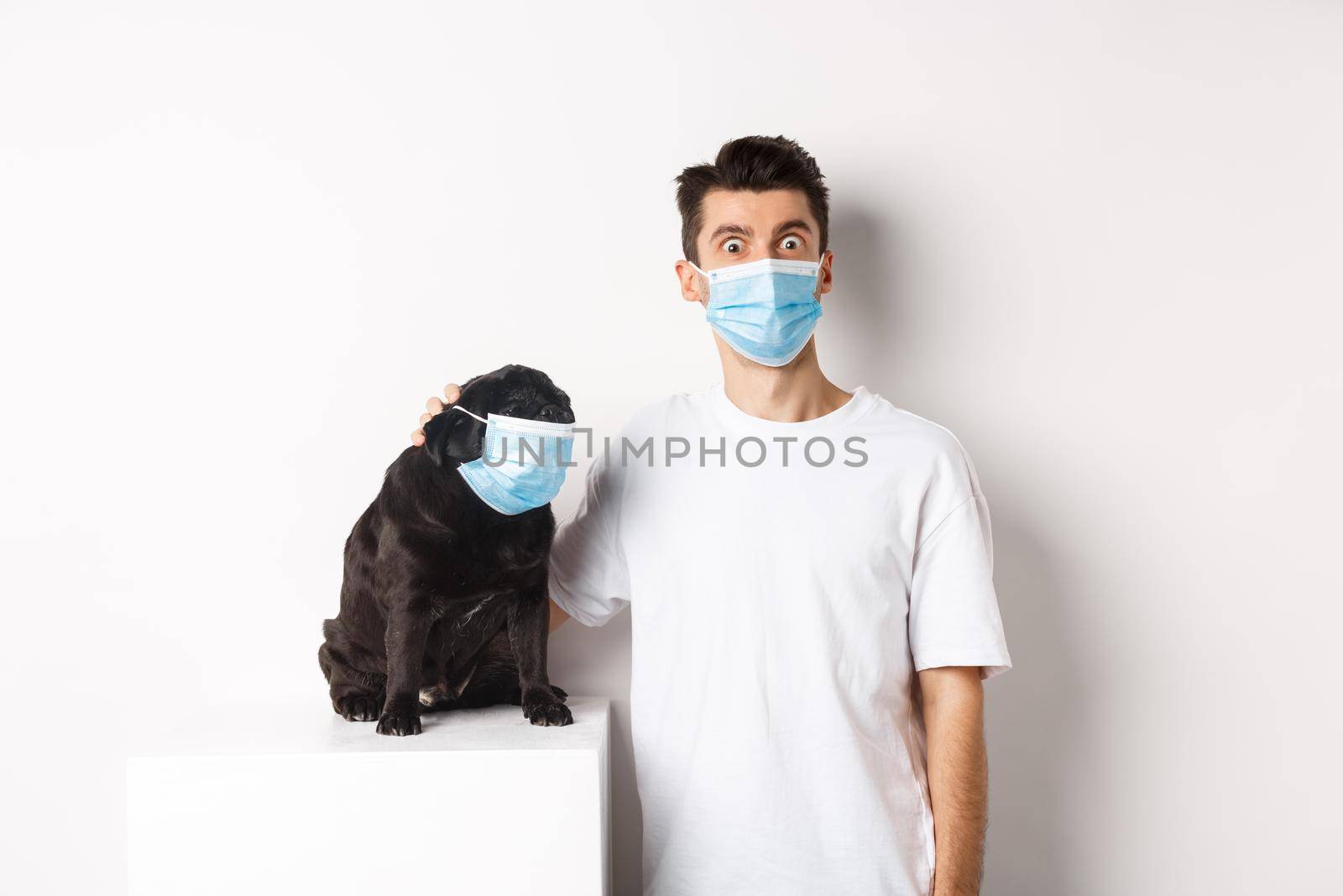 Covid-19, animals and quarantine concept. Image of funny young man and pug in medical masks, staring at camera, standing over white background by Benzoix