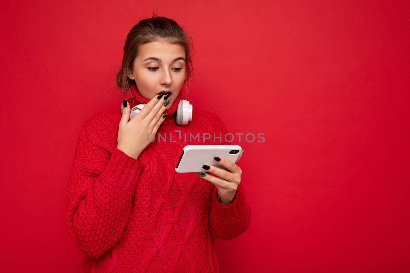 Attractive shocked young brunet woman wearing red sweater isolated over red background wall holding and using mobile phone surfing on the internet and wearing bluetooth headphones looking at smartphone. empty space