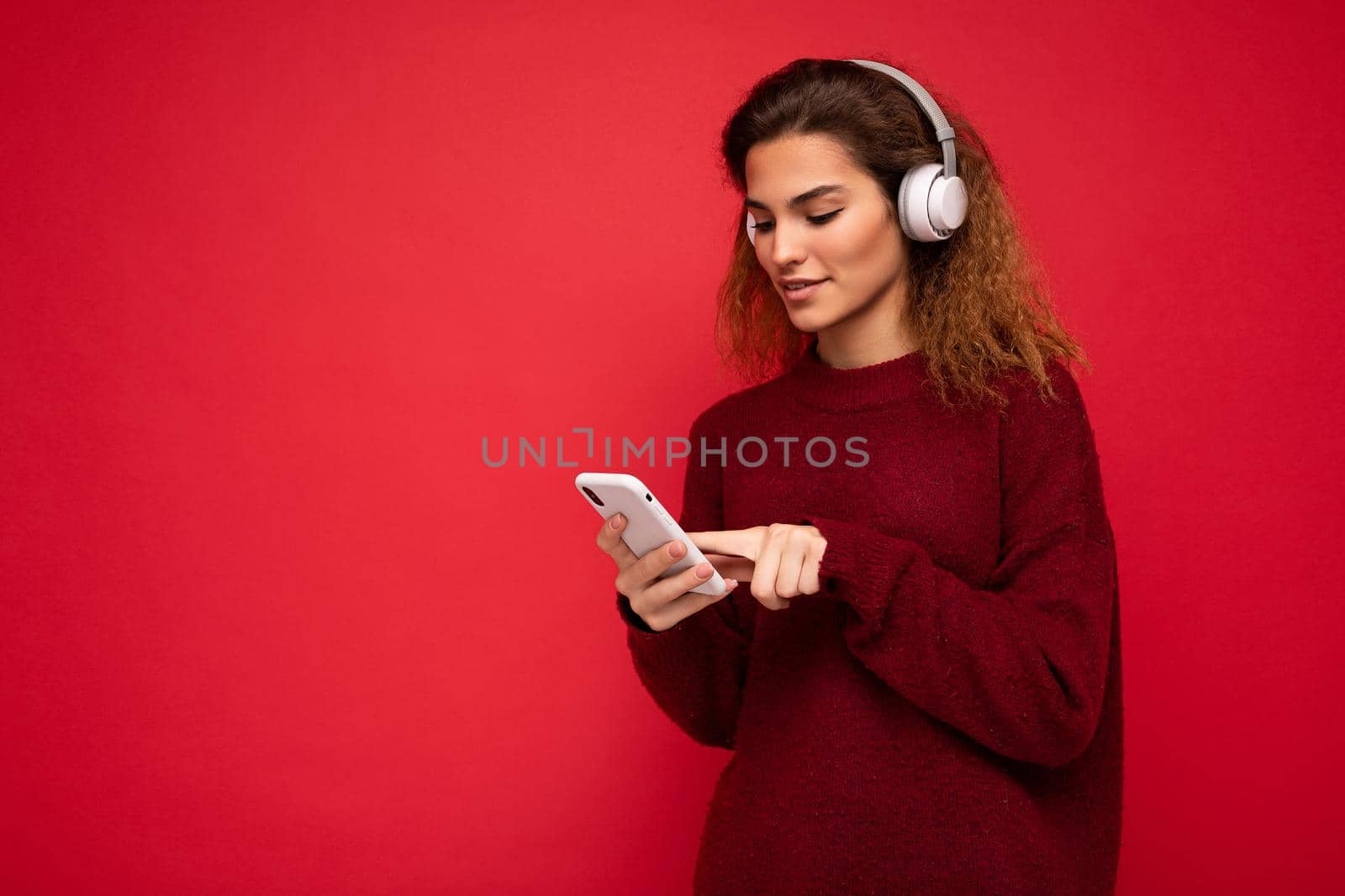 Shot of beautiful young brunette curly woman wearing dark red sweater isolated over red background wall wearing white wireless headphones and listening to music and using mobile phone surfing on the internet looking at smartphone display. copy space
