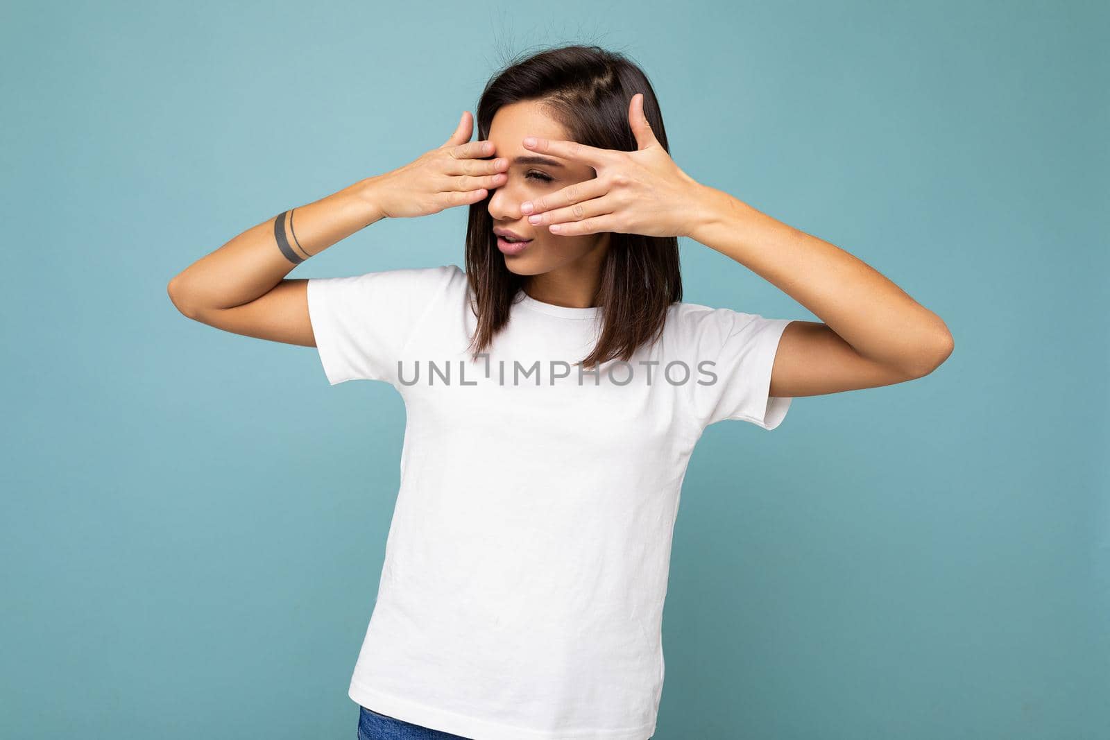 Portrait of young beautiful brunette woman with sincere emotions wearing casual white t-shirt for mockup isolated on blue background with empty space and covering eyes with hands.