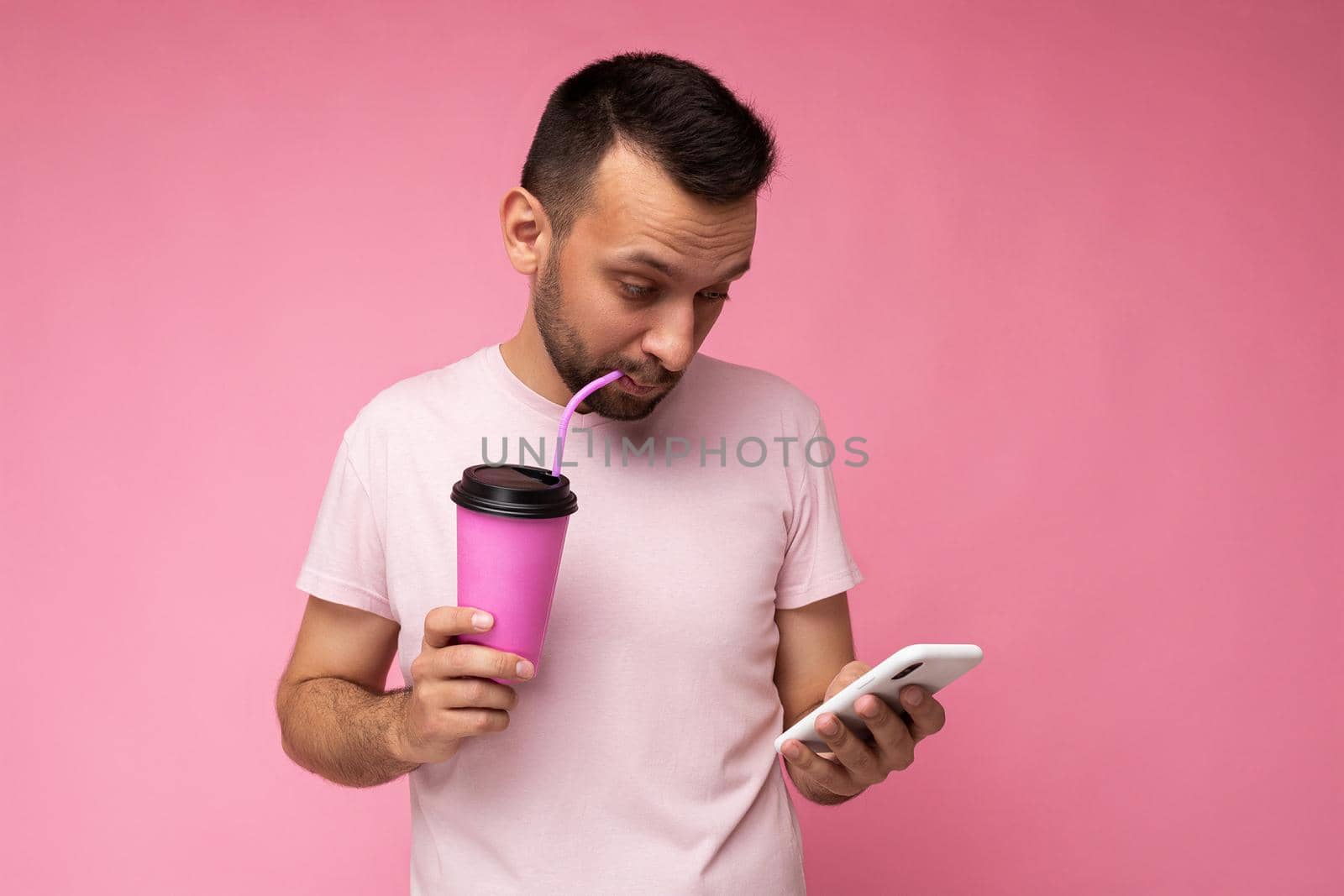 Handsome happy concentrated young brunette unshaven man with beard wearing white t-shirt isolated over pink backgroung wall using mobile phone holding paper coffee cup for mock up and drinking looking at gadjet display. copy space