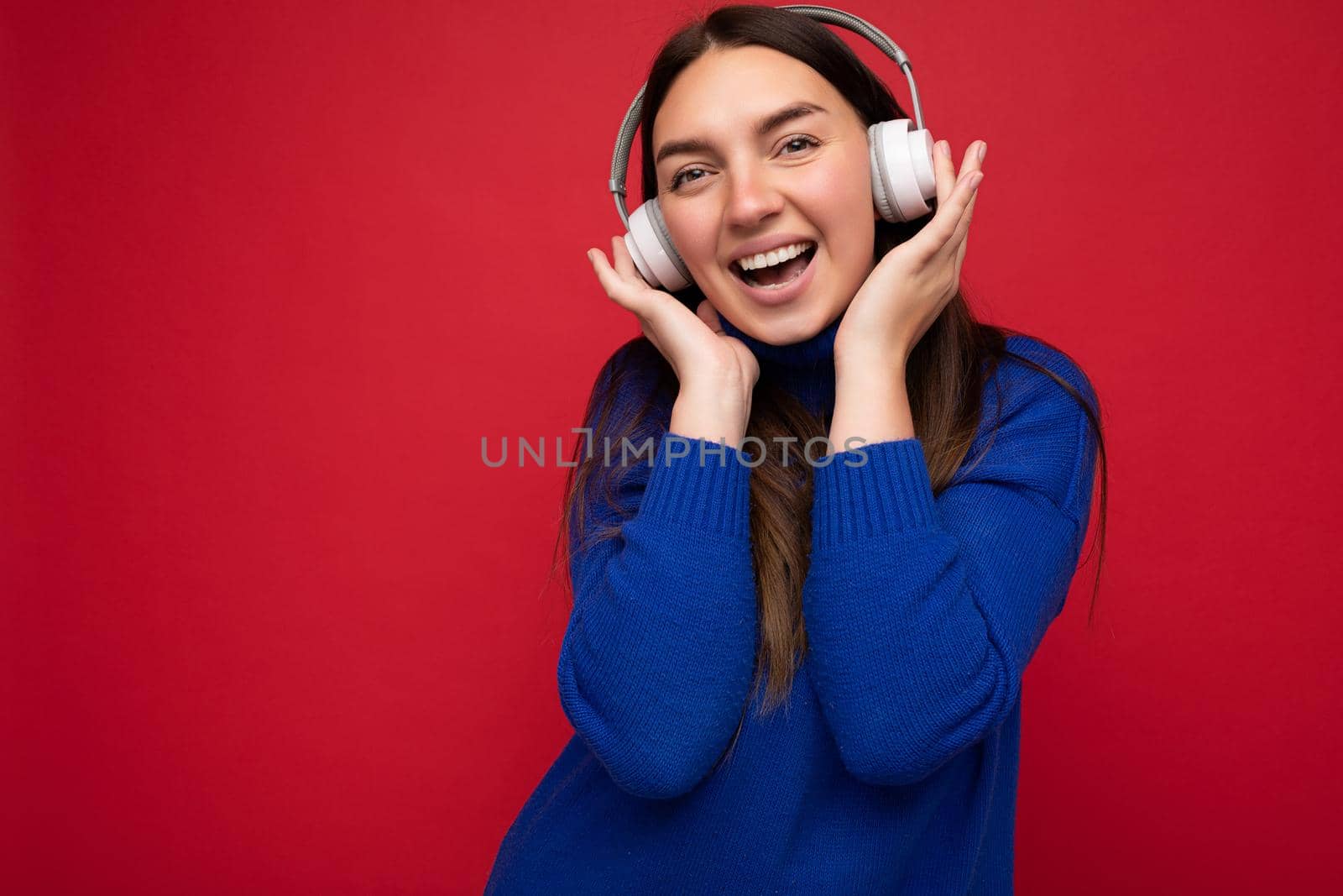 Attractive positive young brunette woman wearing blue sweater isolated over red background wall wearing white bluetooth earphones listening to cool music and having fun looking at camera.