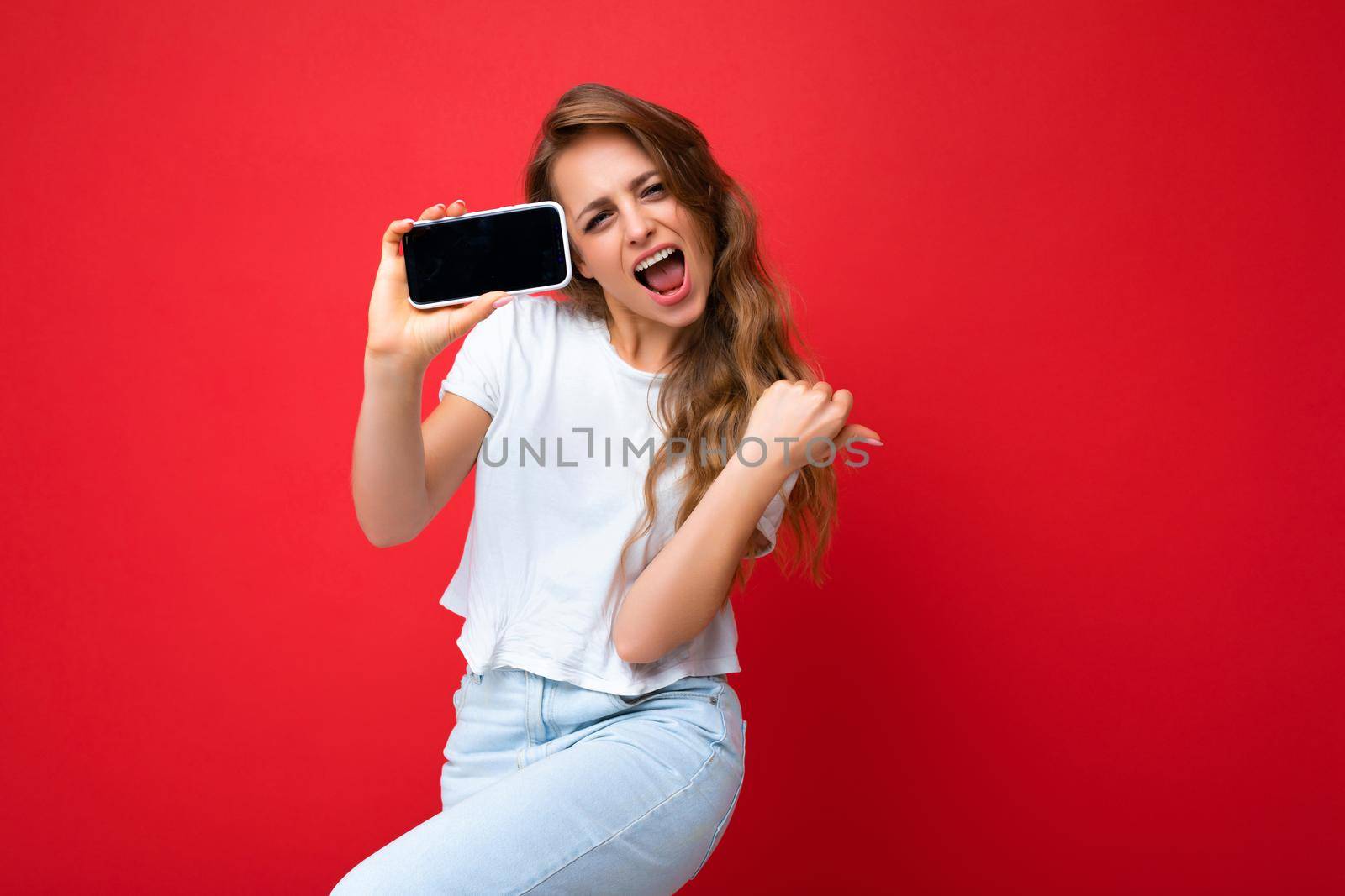Happy satisfied beautiful smiling young blonde woman good looking wearing white t-shirt standing isolated on red background with copy space holding phone showing smartphone in hand with empty screen display for mockup looking at camera.