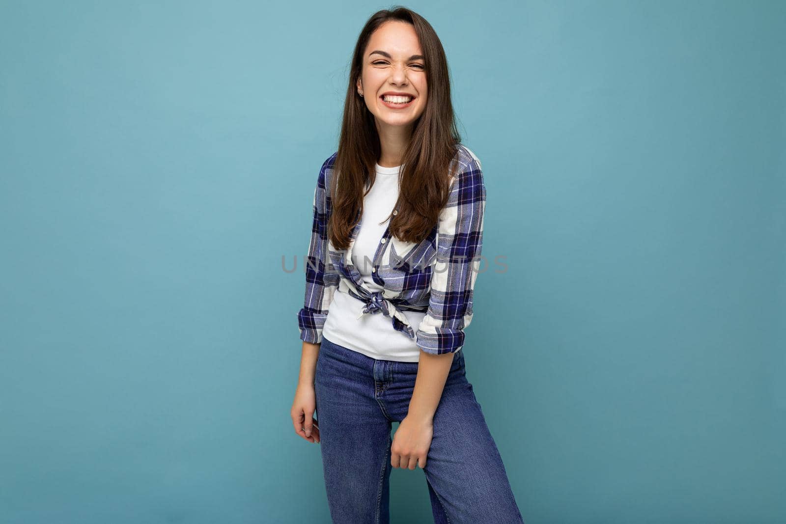 Photo portrait of young beautiful smiling hipster brunette woman in trendy blue and white shirt and jeans clothes. Sexy carefree female person posing isolated near blue wall with empty space in studio. Positive model with natural makeup.