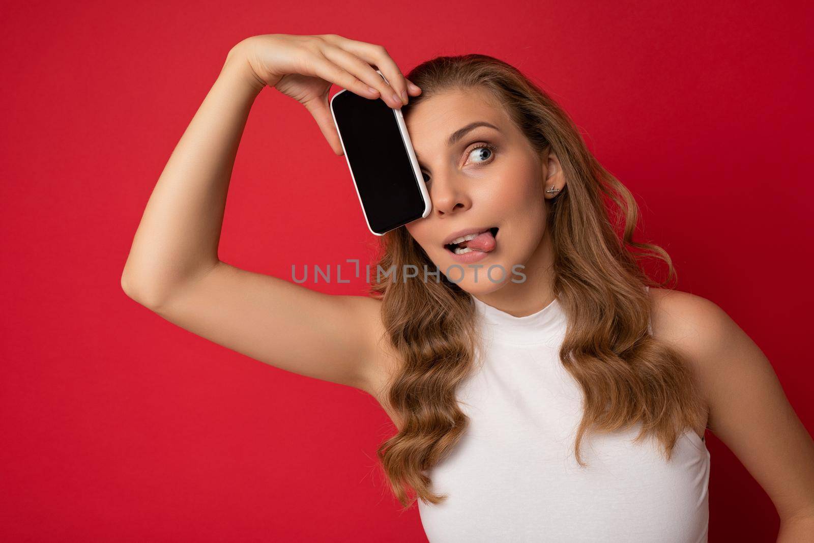 Happy sexy attractive young blonde female person wearing white t-shirt isolated on red background with copy space holding smartphone showing phone in hand with empty screen for cutout looking to the side and showing tongue.