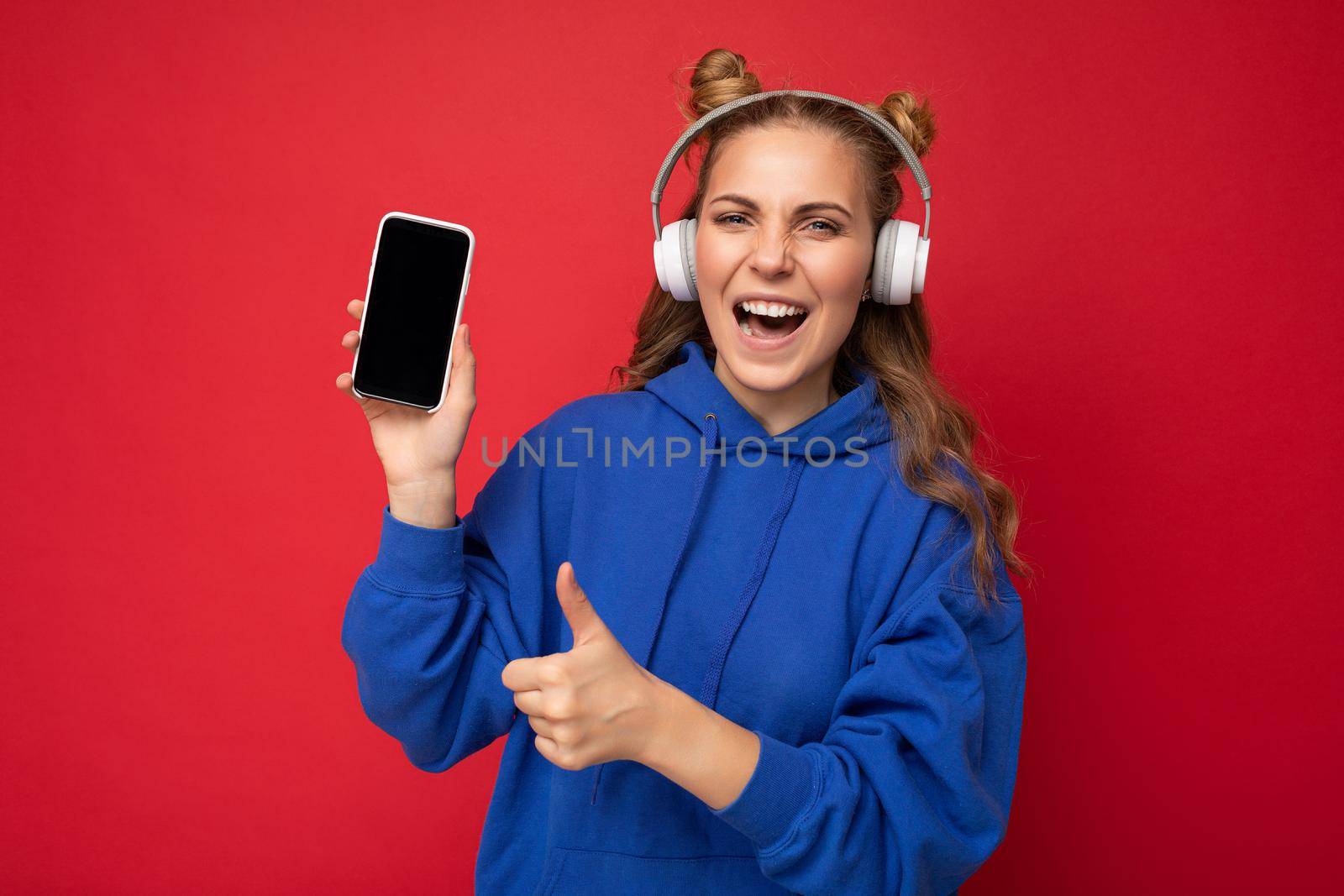 Beautiful happy smiling young woman wearing stylish casual outfit isolated on background wall holding and showing mobile phone with empty display for mockup wearing white bluetooth headphones listening to music and having fun looking at camera and showing thumbs up gesture.