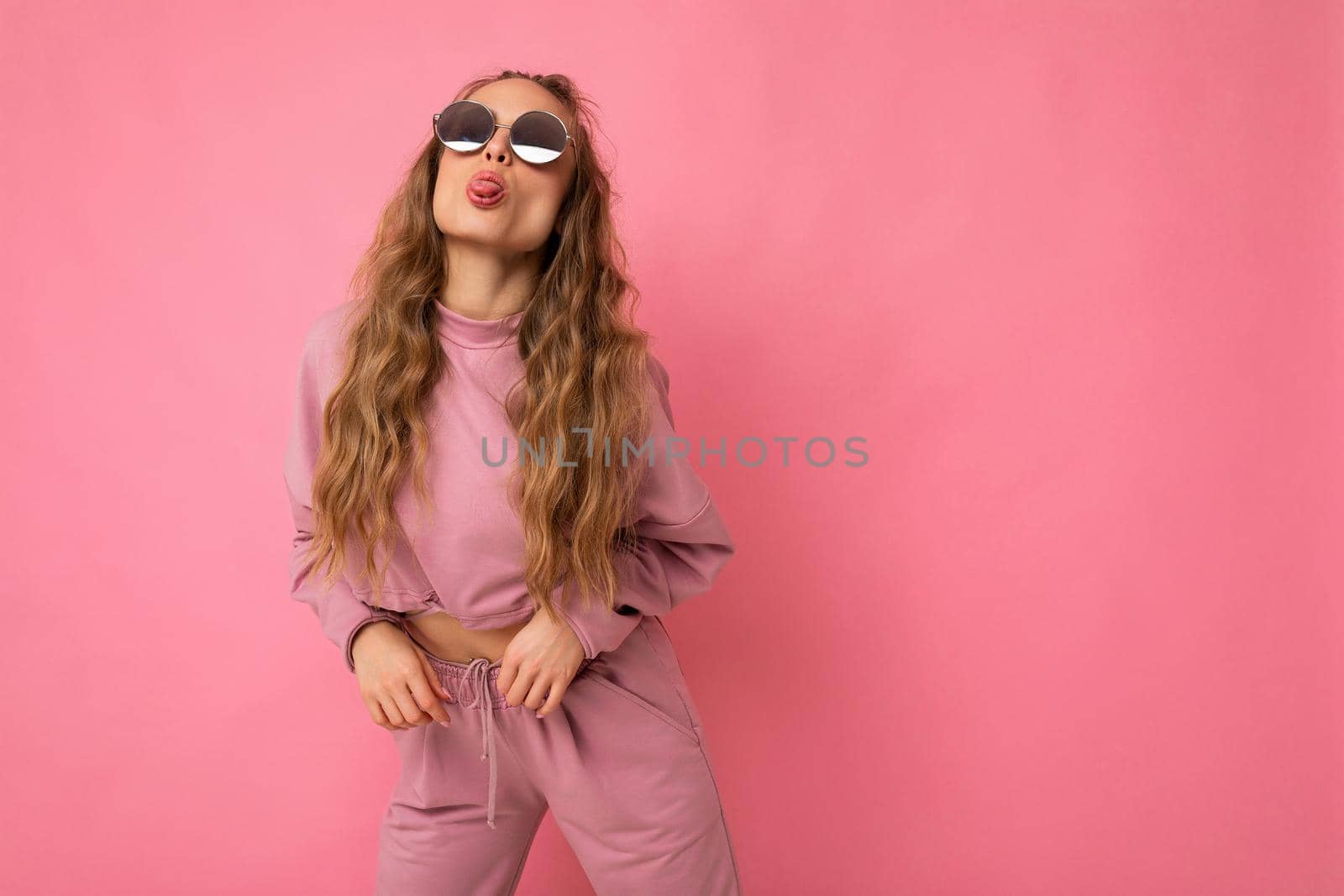 Shot of fascinating positive happy young dark blonde curly woman isolated over pink background wall wearing casual pink sport clothes and stylish sunglasses and having fun. empty space